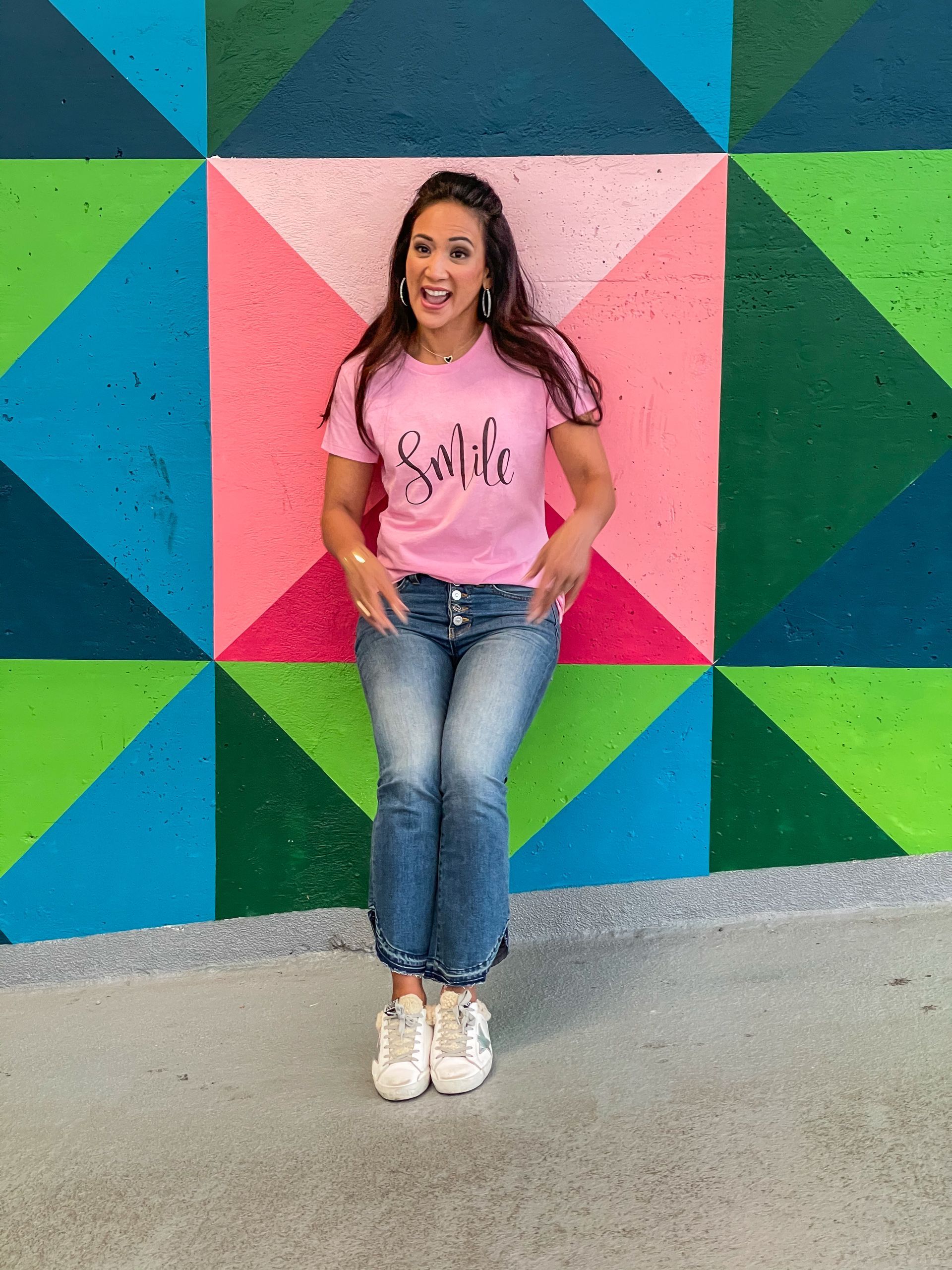 Woman in pink shirt and jeans smiles in front of a colorful geometric wall.