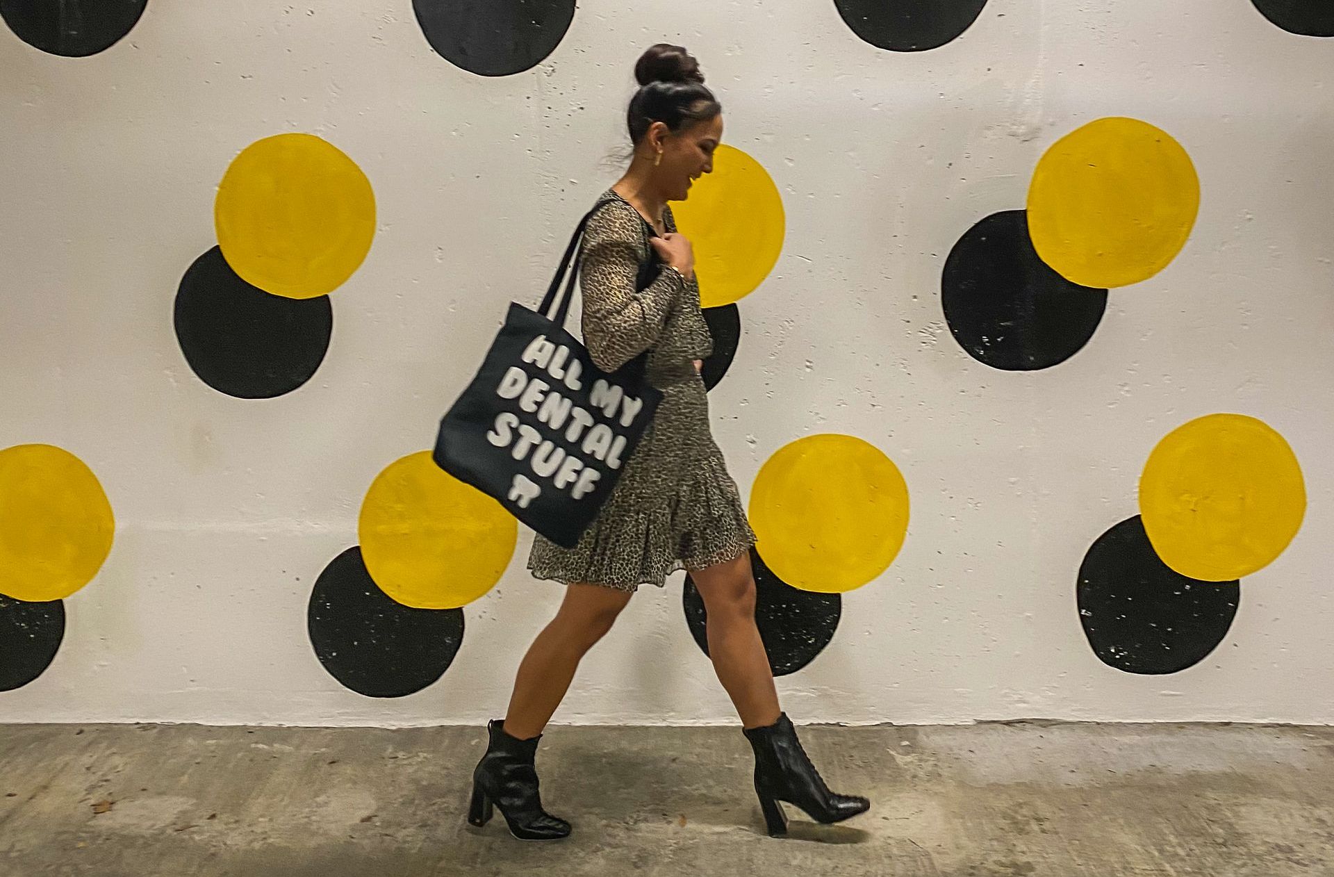 Woman in a dress walks past a polka dot wall, carrying a tote bag that says 