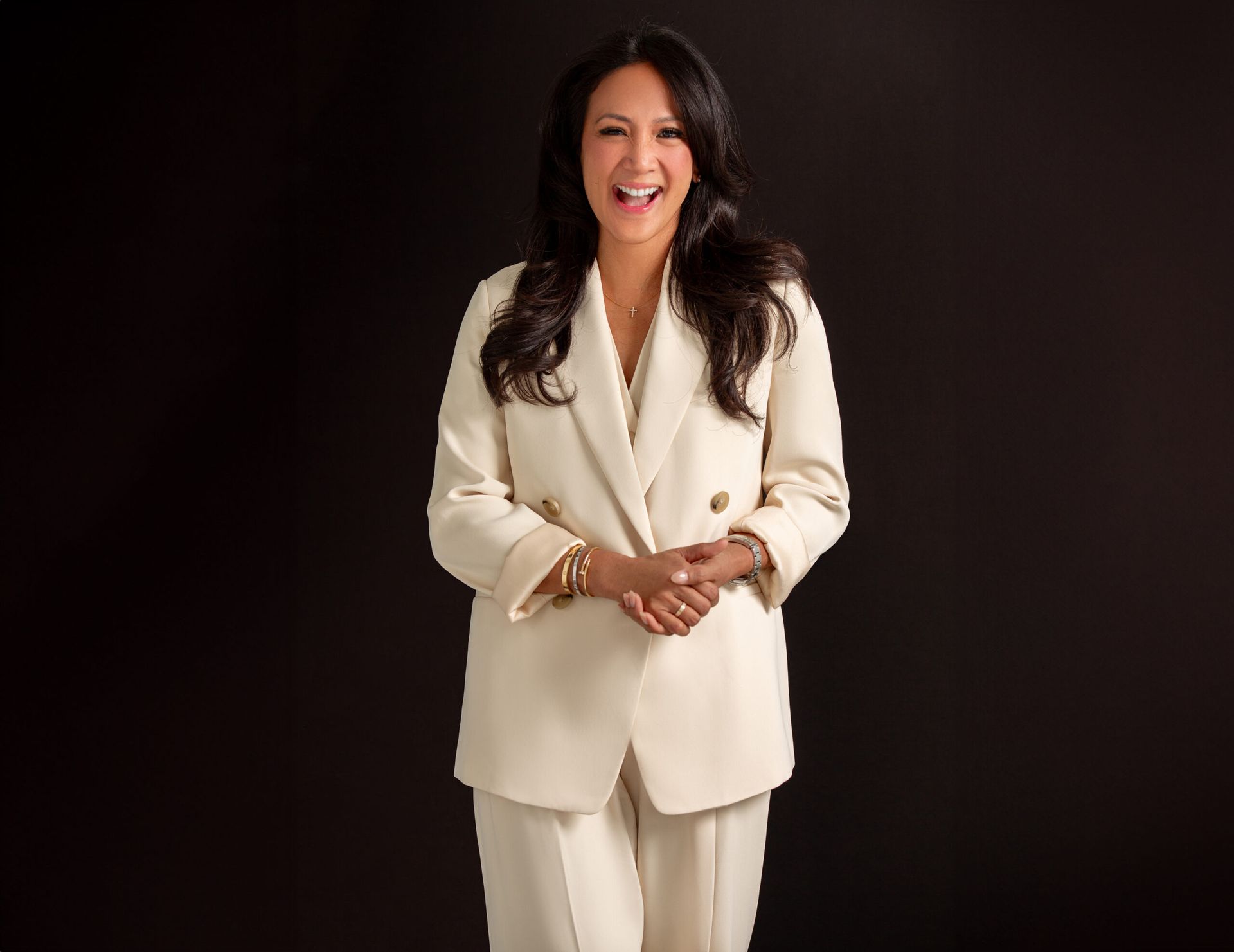 Woman in cream suit, smiling widely, hands clasped, against a black backdrop.