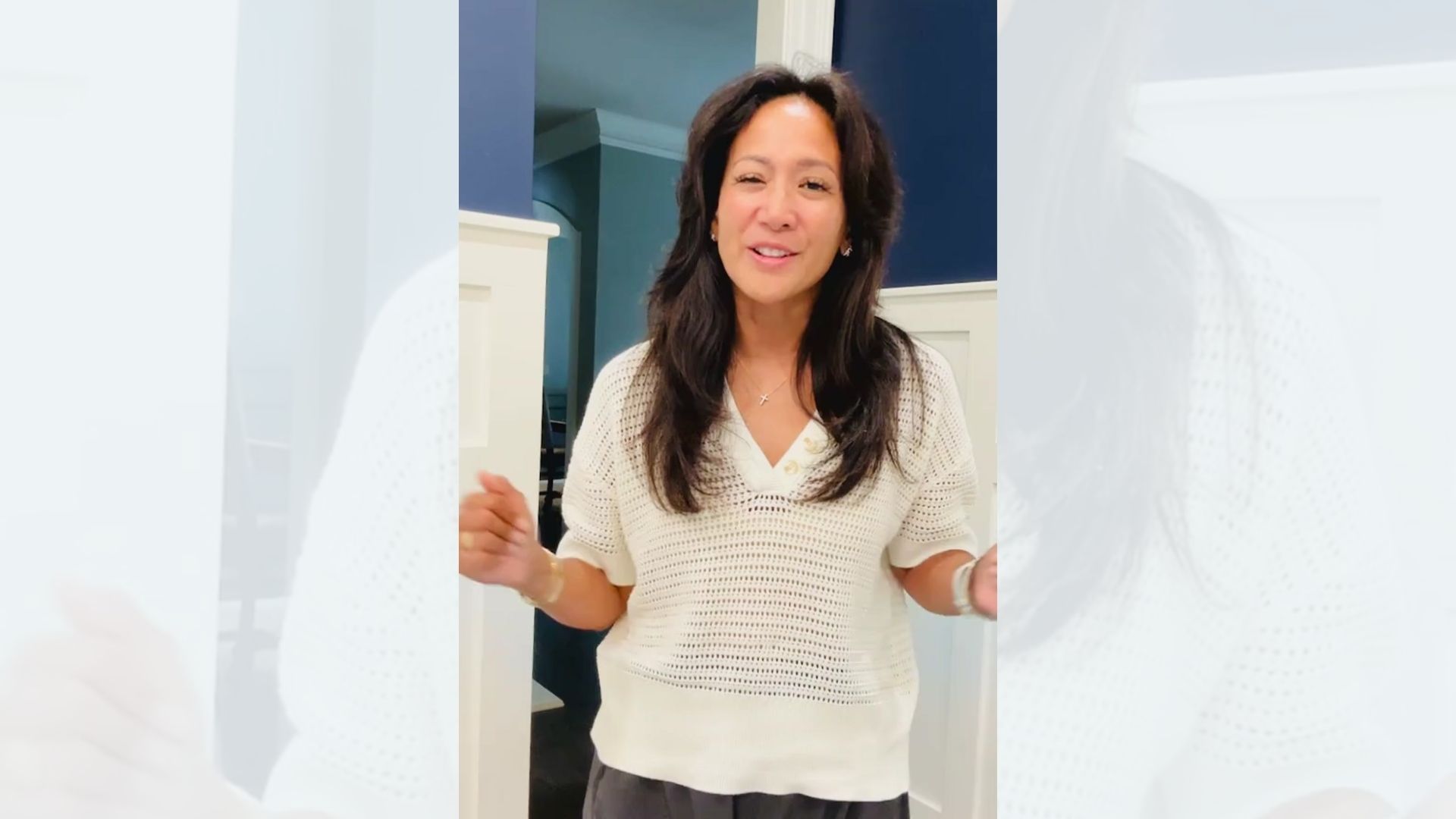 Woman with dark hair, wearing a cream sweater, standing in a room, smiling and gesturing.