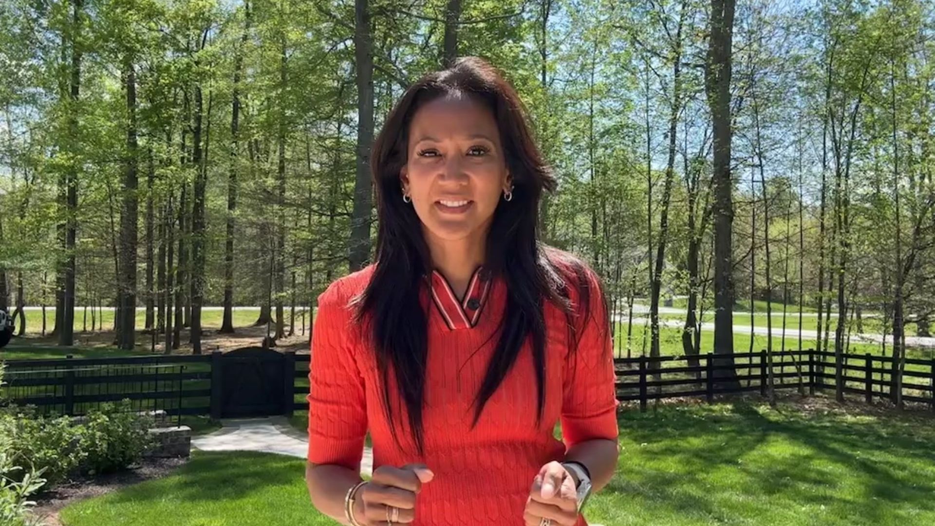 Woman in orange top smiles in front of trees, a fence, and clear blue sky.