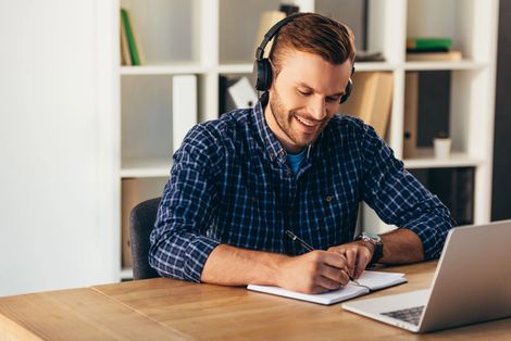 Hombre con auriculares escribiendo en un cuaderno junto a un portátil en un escritorio en una oficina luminosa.