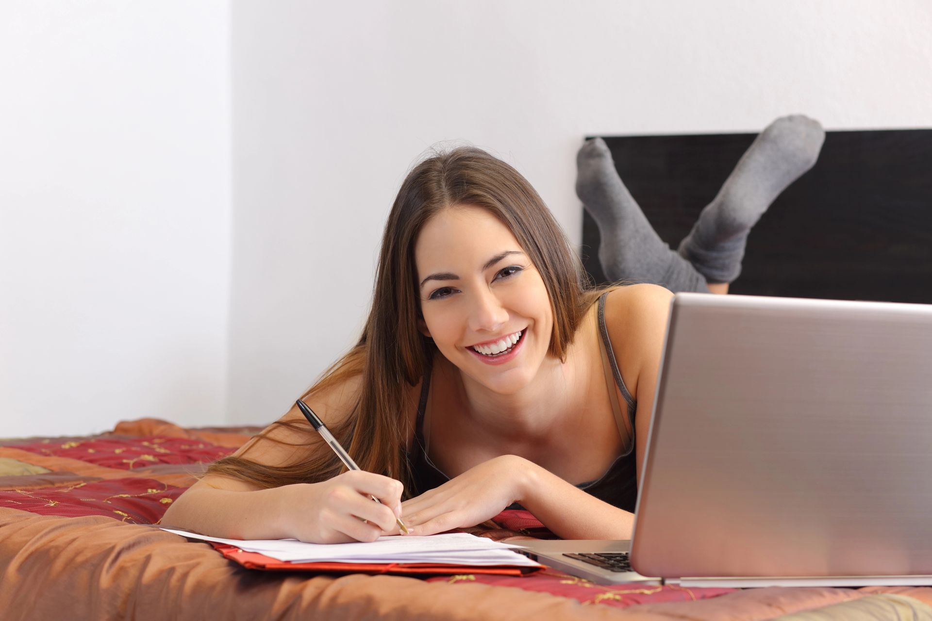 Mujer sonriente escribiendo en un cuaderno junto a una computadora portátil en una cama.