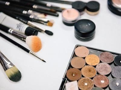 Pennelli per il trucco, palette di ombretti e contenitori per cosmetici su una superficie bianca.