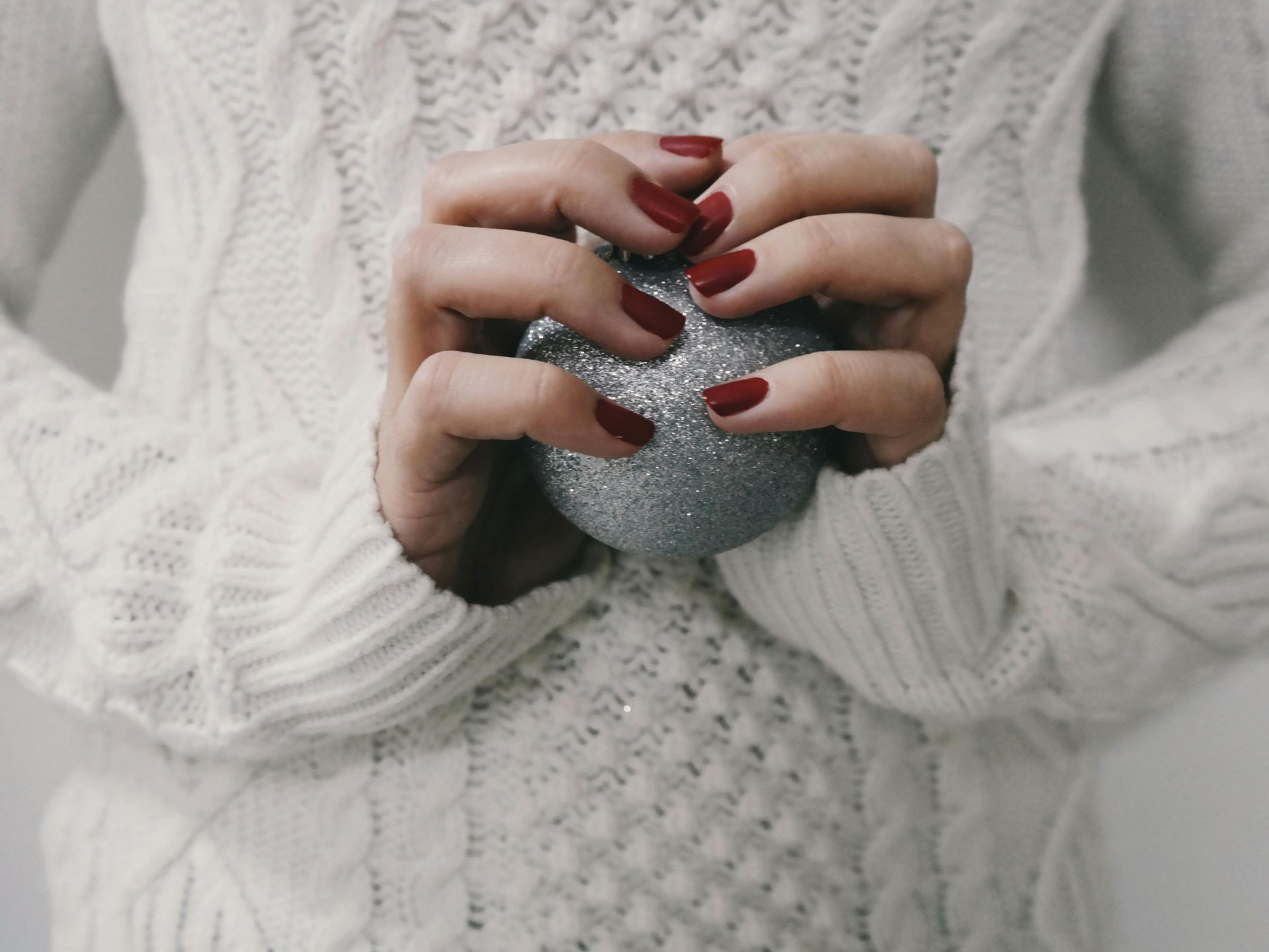 Mani con smalto rosso che tengono un ornamento glitterato argentato, indossa un maglione bianco lavorato a maglia.