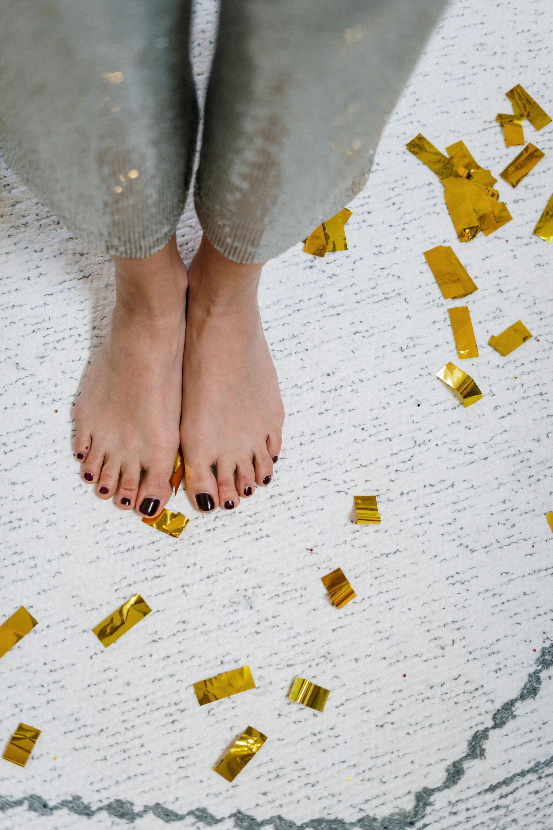 Piedi nudi con smalto rosso scuro su un tappeto bianco con coriandoli dorati, pantaloni con paillettes argentate sullo sfondo.