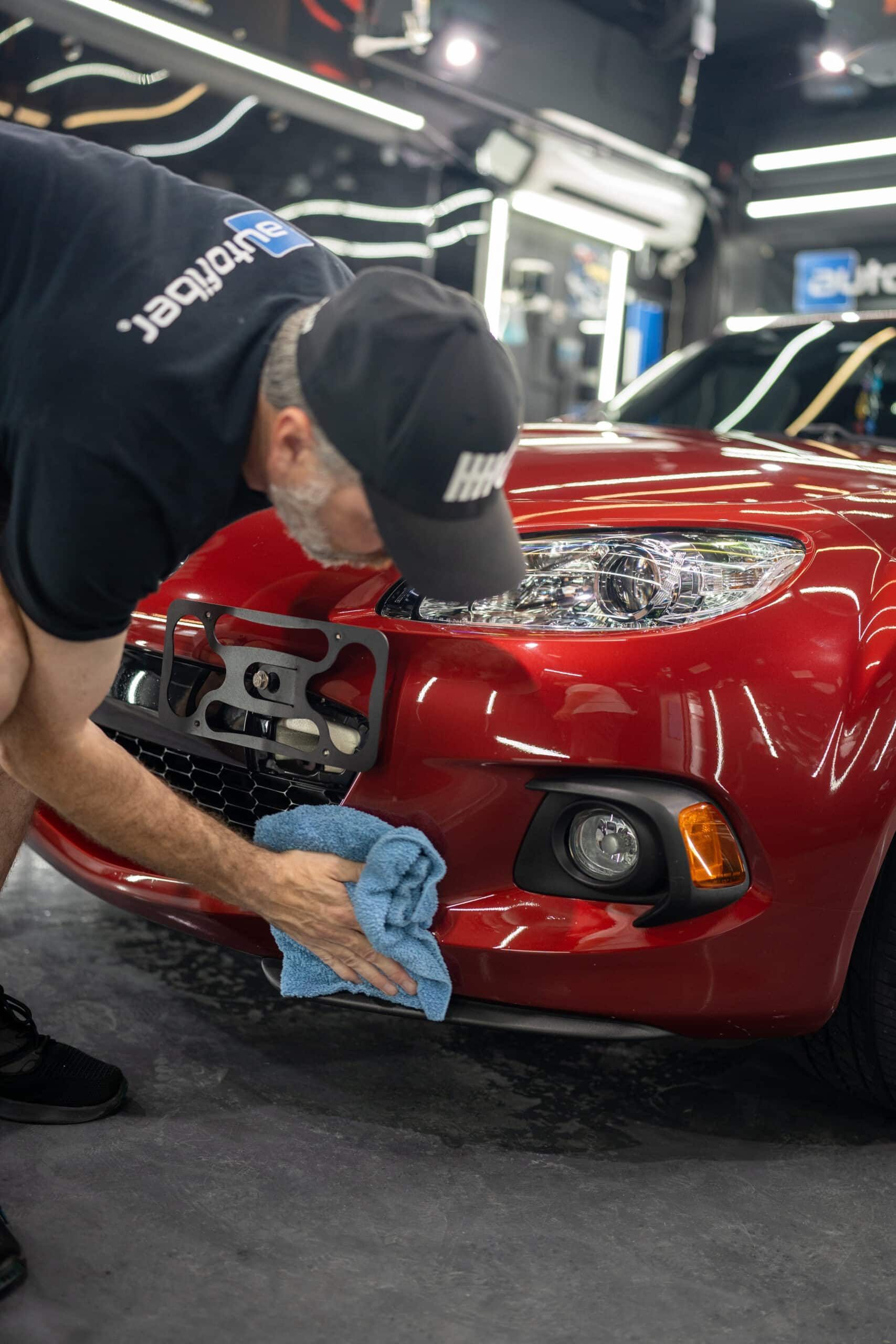A man is cleaning the front of a red car with a towel.