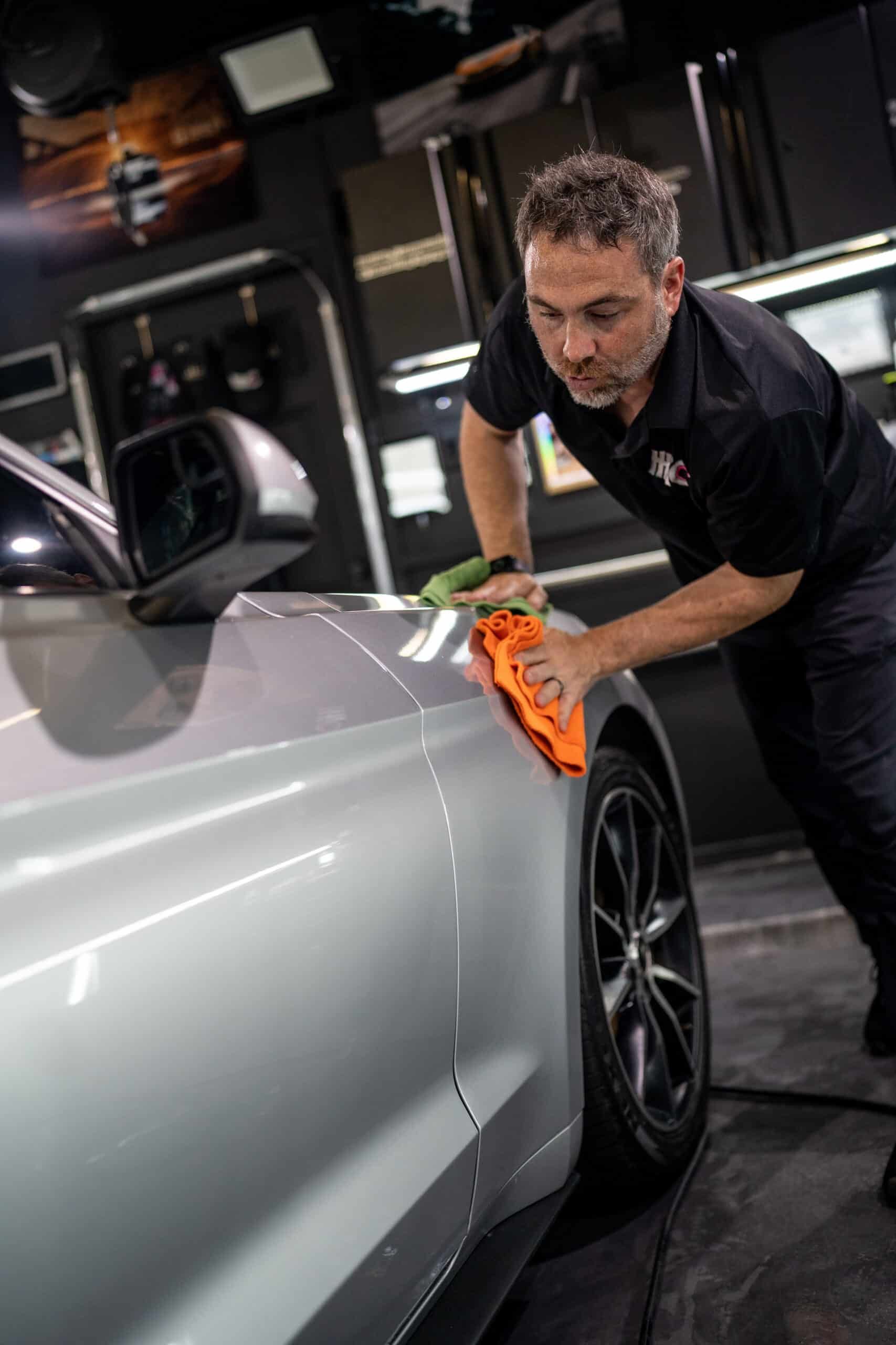 A man is polishing a silver car with an orange cloth.