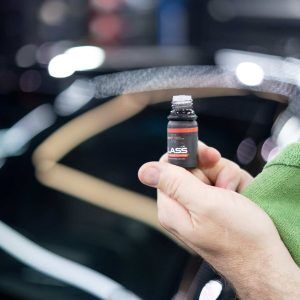 A person is holding a small bottle of liquid in front of a car windshield.