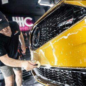 A man is cleaning the front bumper of a yellow car.