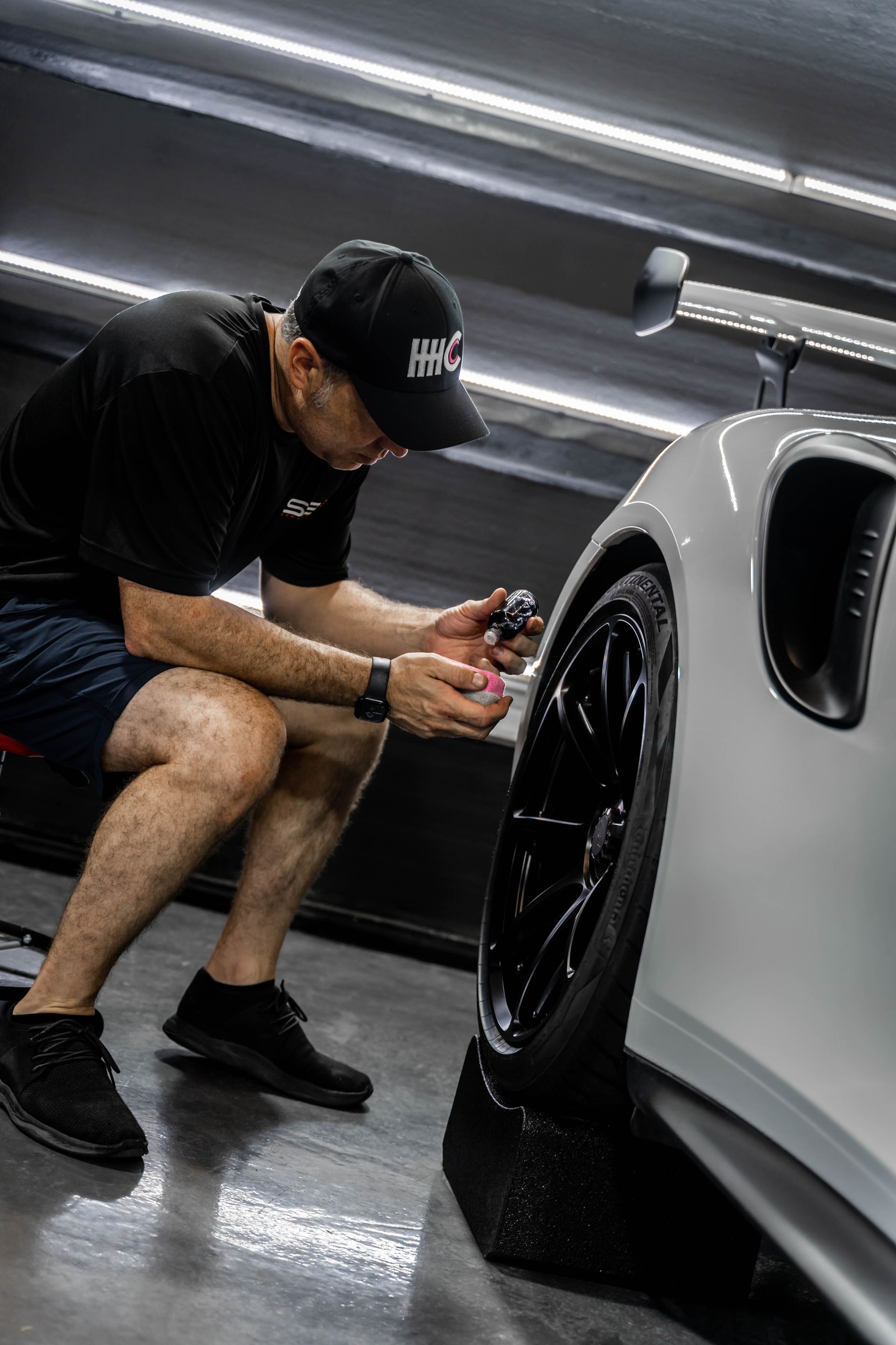 A man is kneeling down next to a white car in a garage.