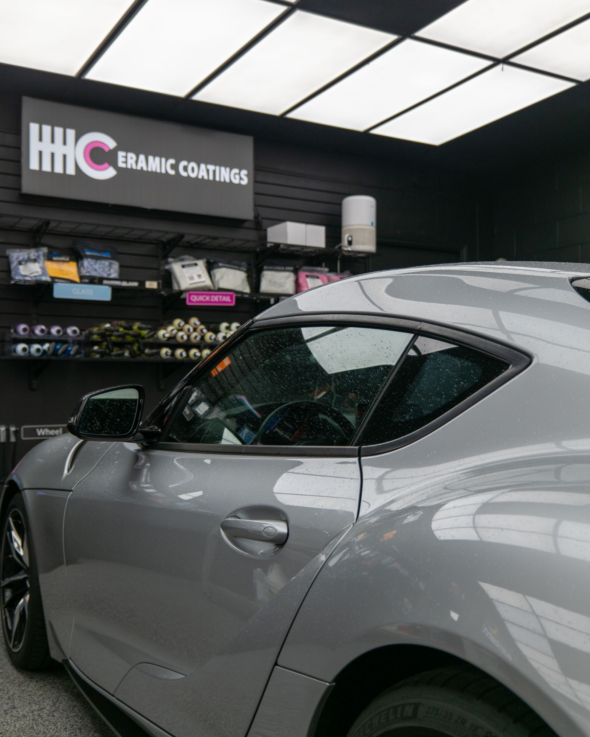 Silver sports coupe in a ceramic coating shop, with shelves and bright ceiling lights in the background