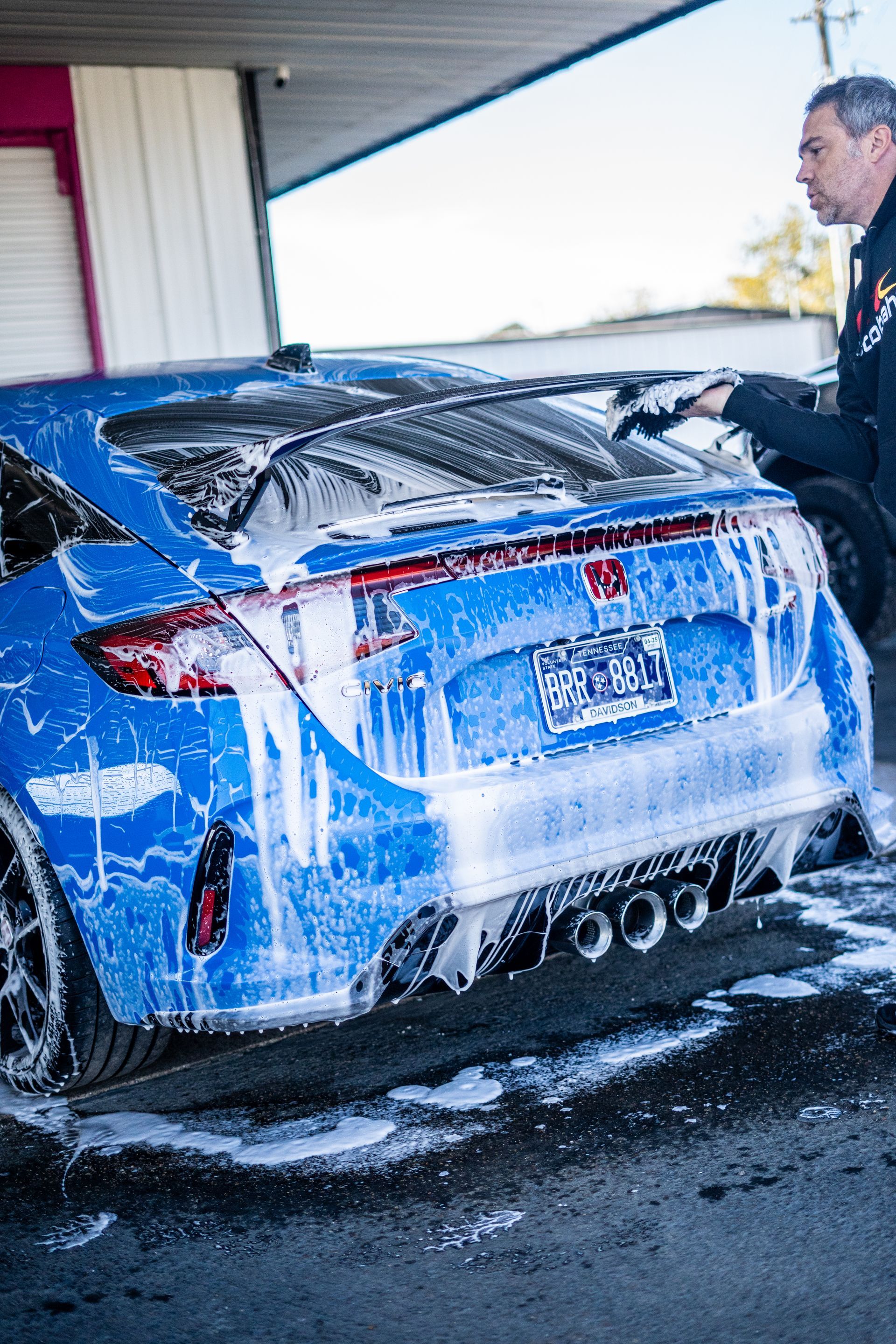 Blue car being washed with foamy soap by a person in an outdoor setting.