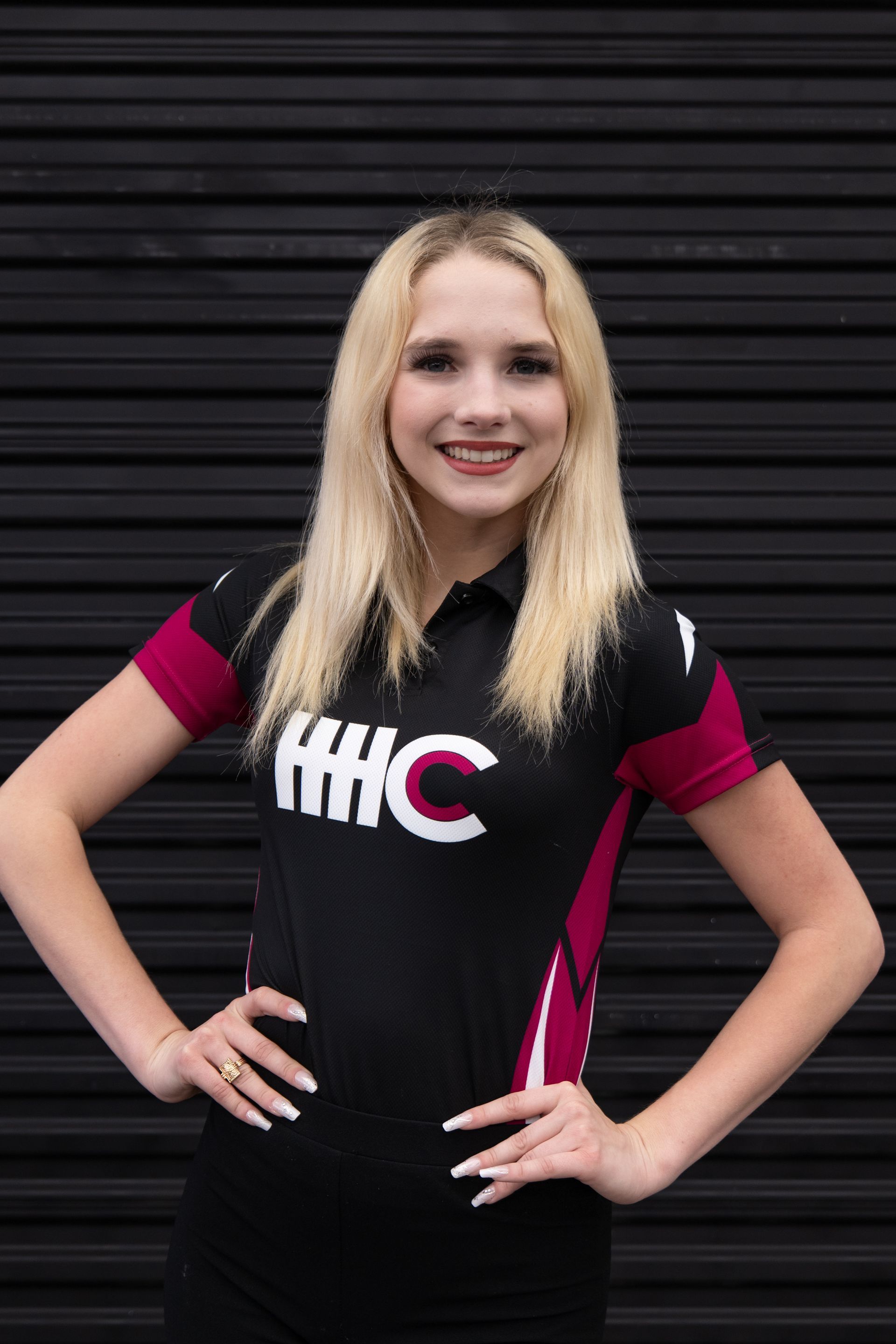 A smiling person with blonde hair wears a black and maroon HHC sports polo, posing with hands on hips against a black wall.