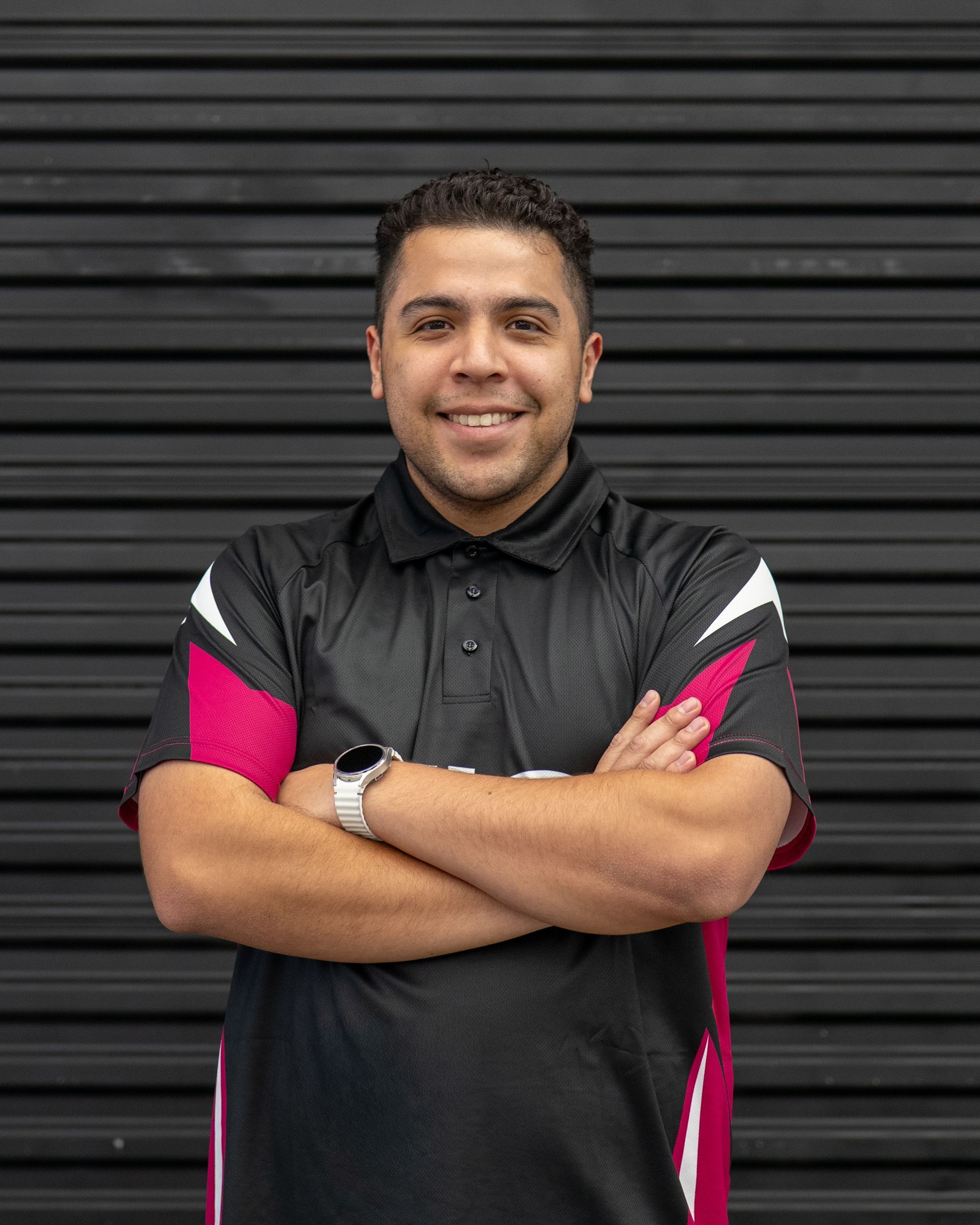 Person in a black and pink polo shirt, smiling with arms crossed in front of a dark shutter.