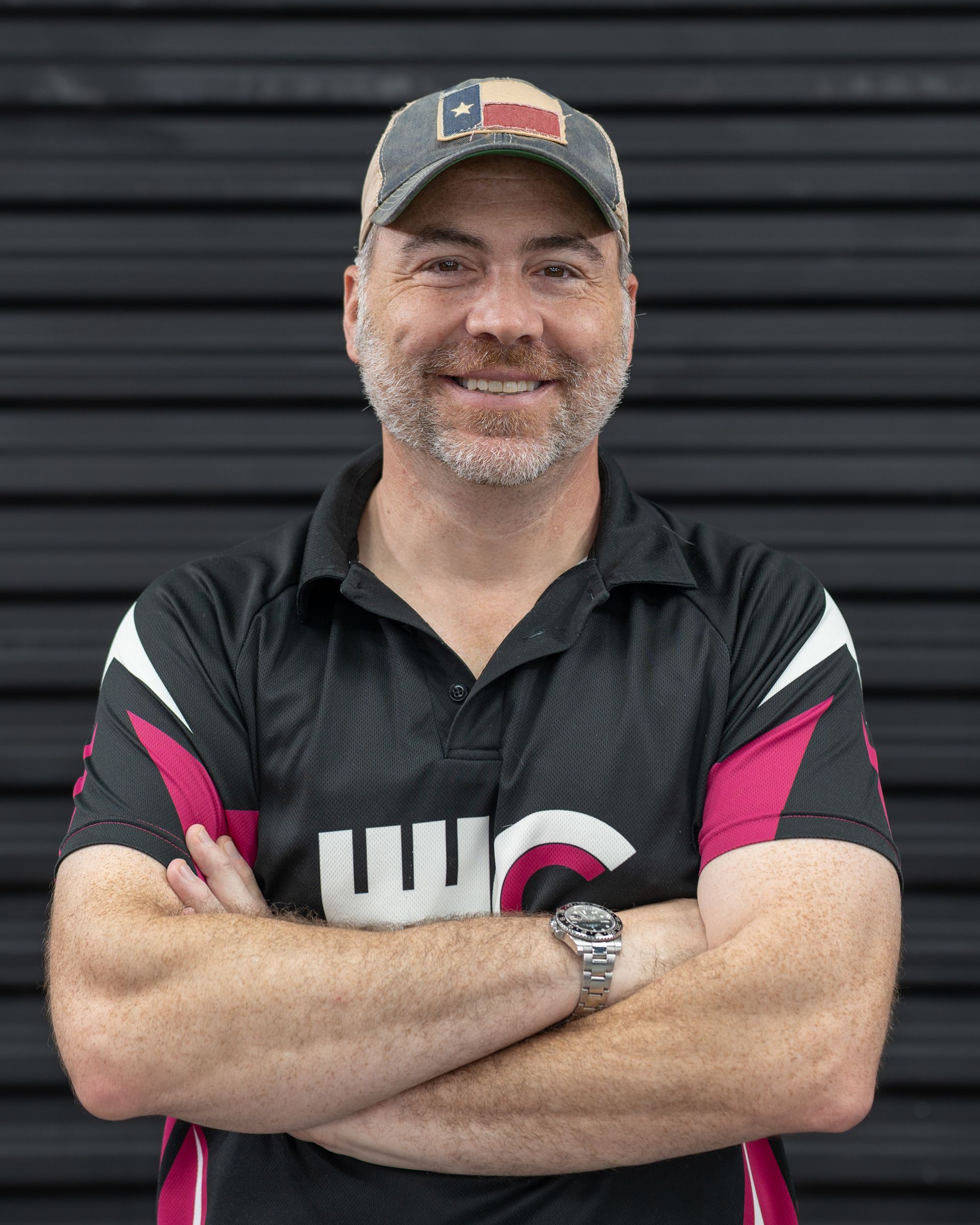Man in Texas hat, arms crossed, smiling. Wearing a black and pink shirt, in front of a dark corrugated wall.