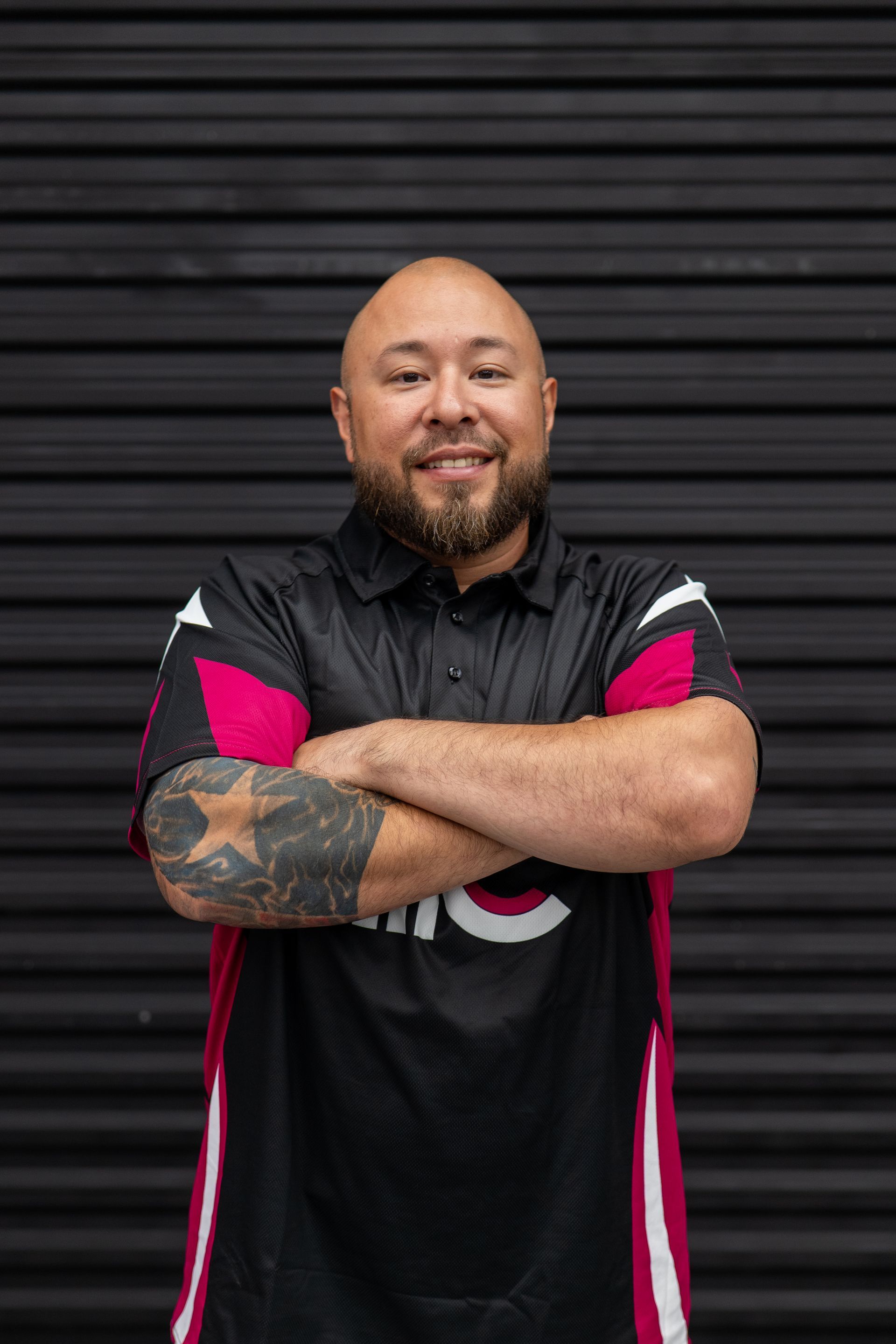 Man with tattoos and crossed arms, wearing a black shirt with pink accents in front of a dark background.