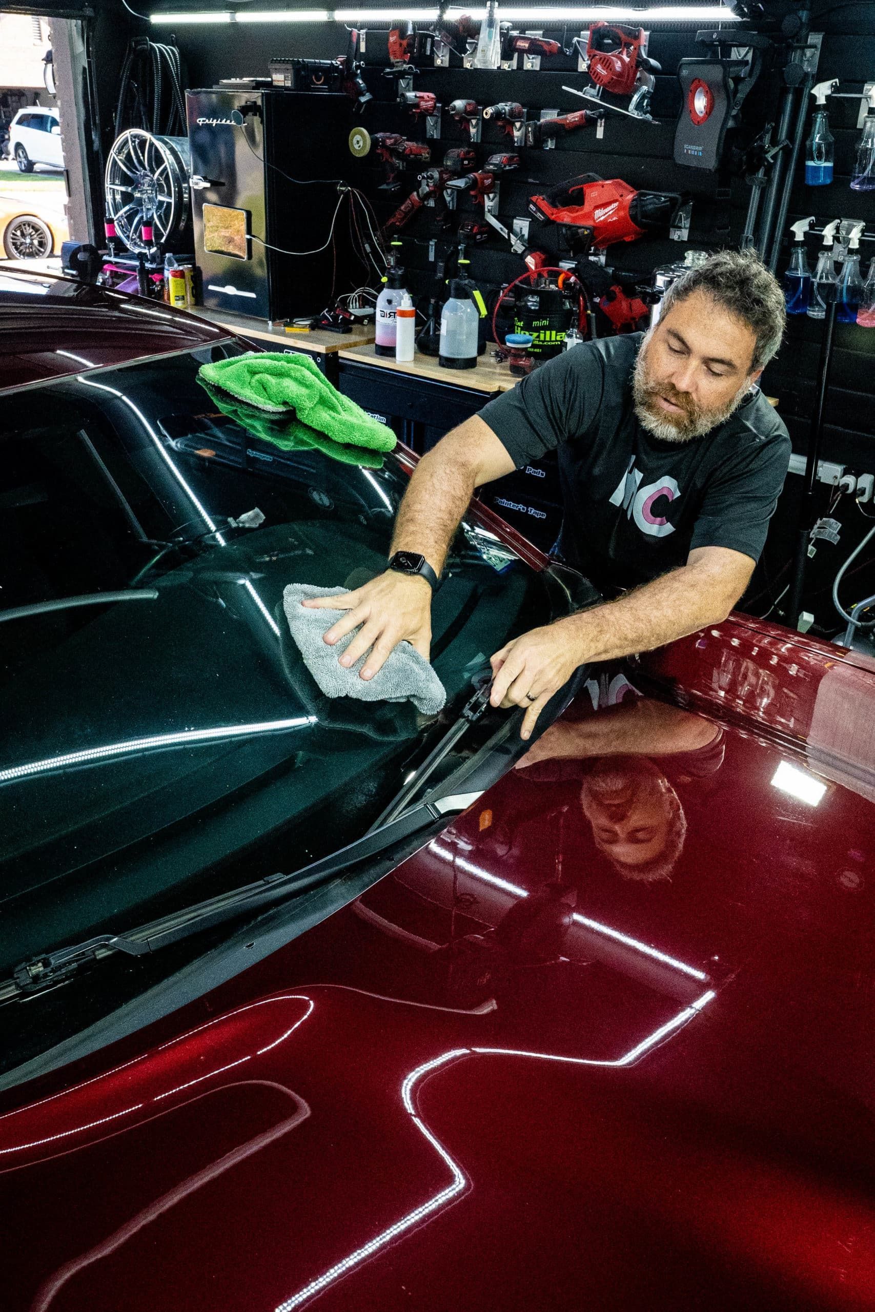A man is cleaning the windshield of a red car in a garage.