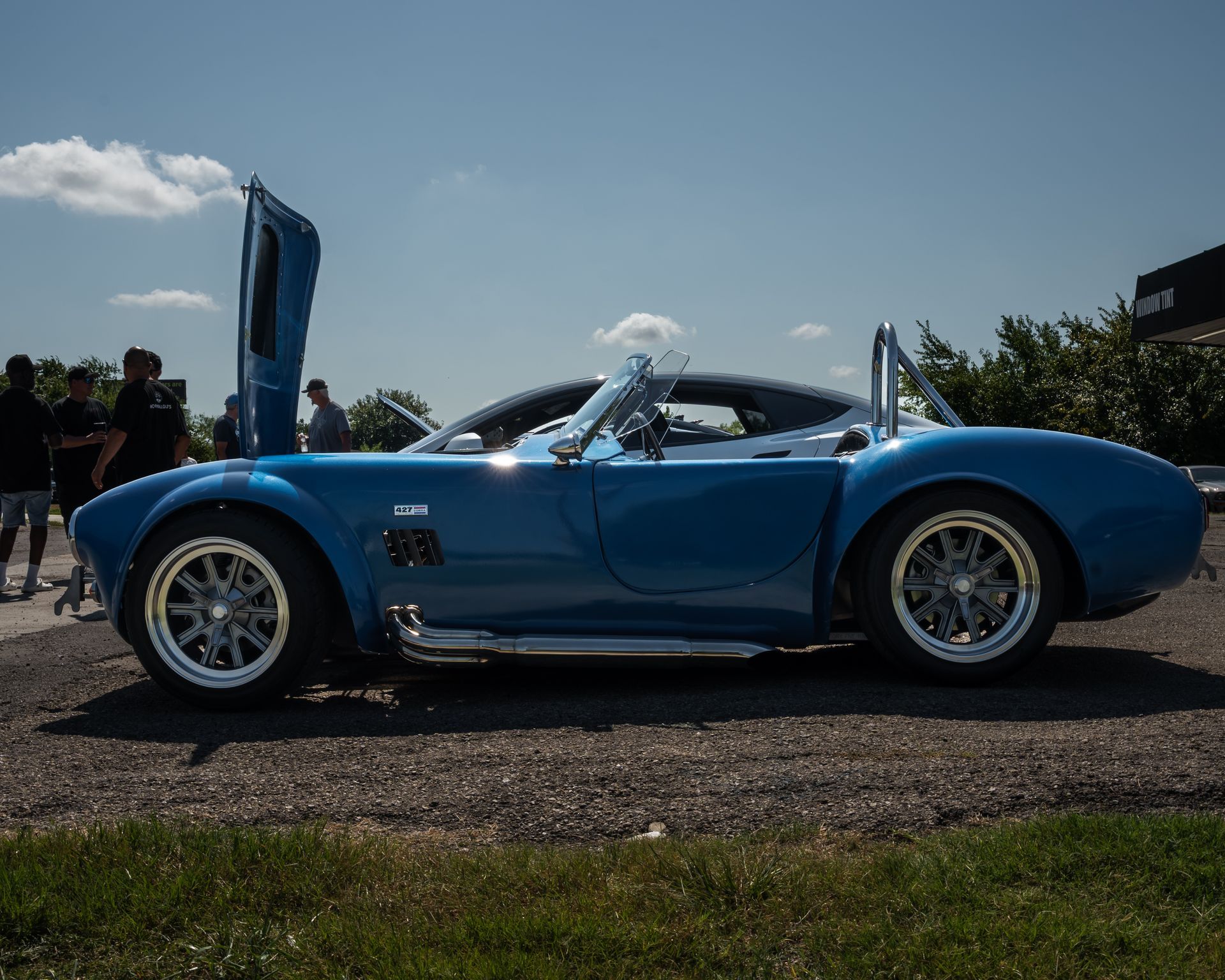 Blue classic sports car with doors open, parked on gravel, under a blue sky.