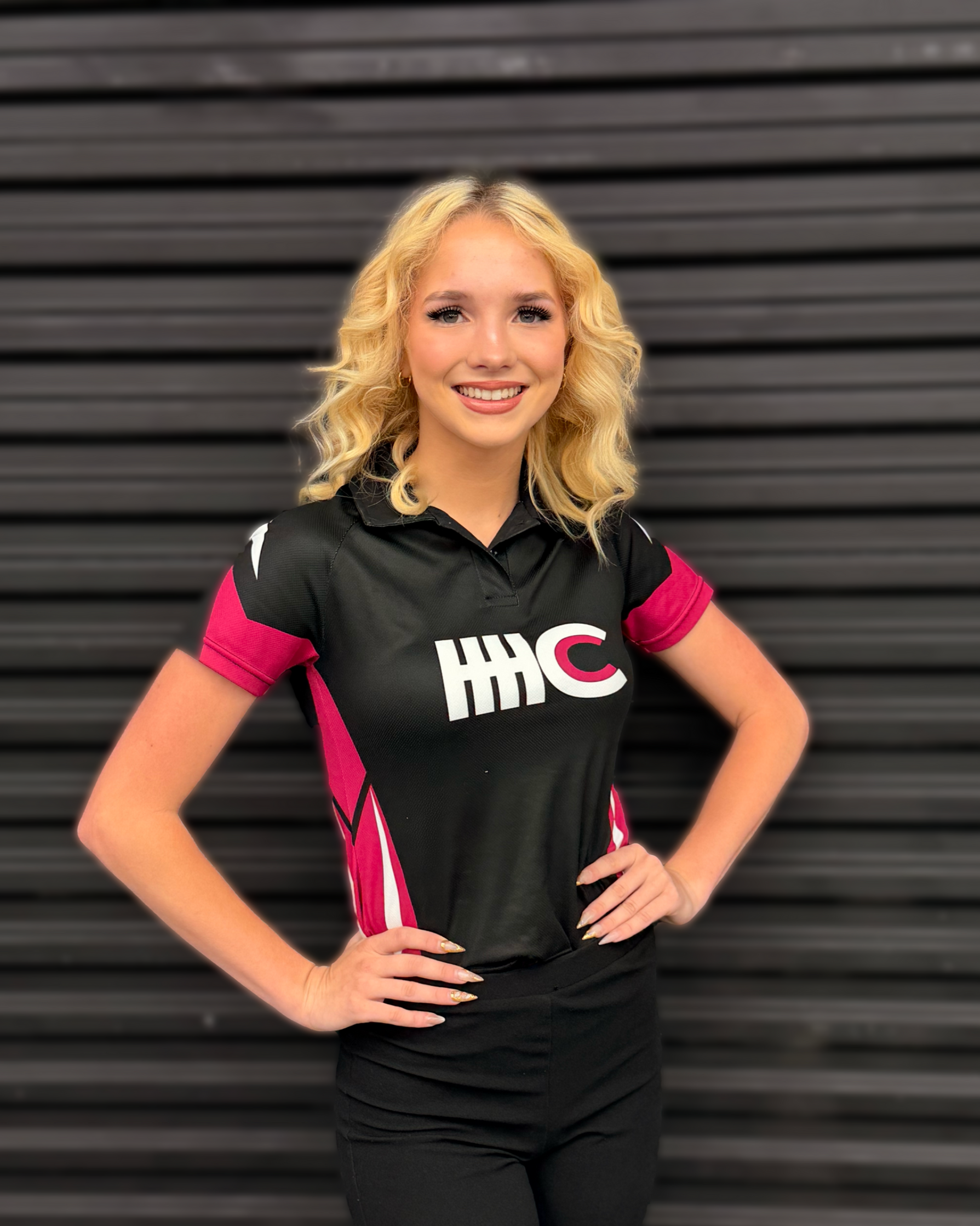 A smiling person with blonde hair wears a black and maroon HHC sports polo, posing with hands on hips against a black wall.