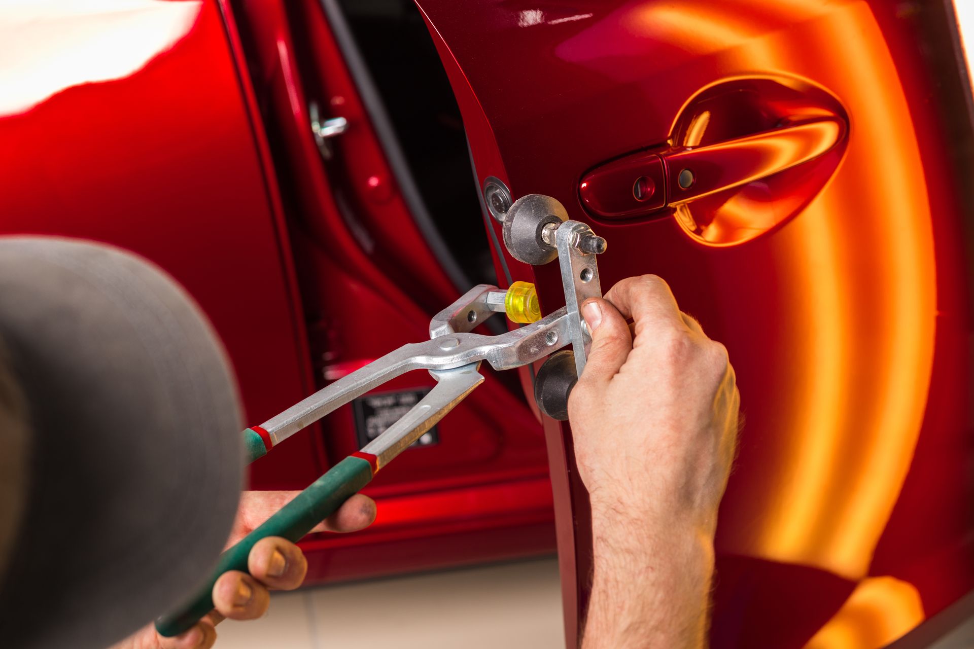 Person using tools to remove a dent from a red car door in a well-lit shop.