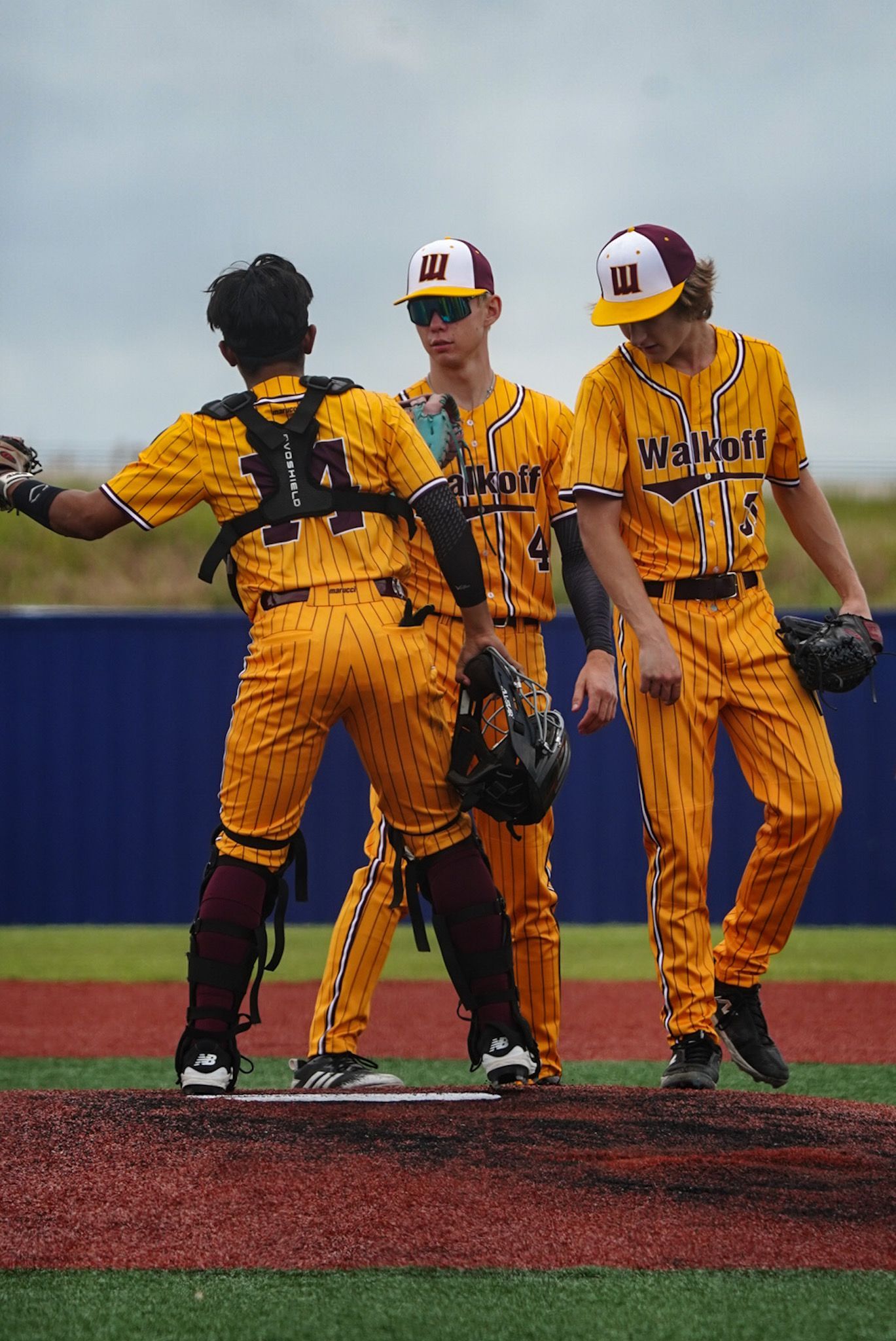 Three softball players in yellow uniforms huddle on a field, with one facing away and others facing her.