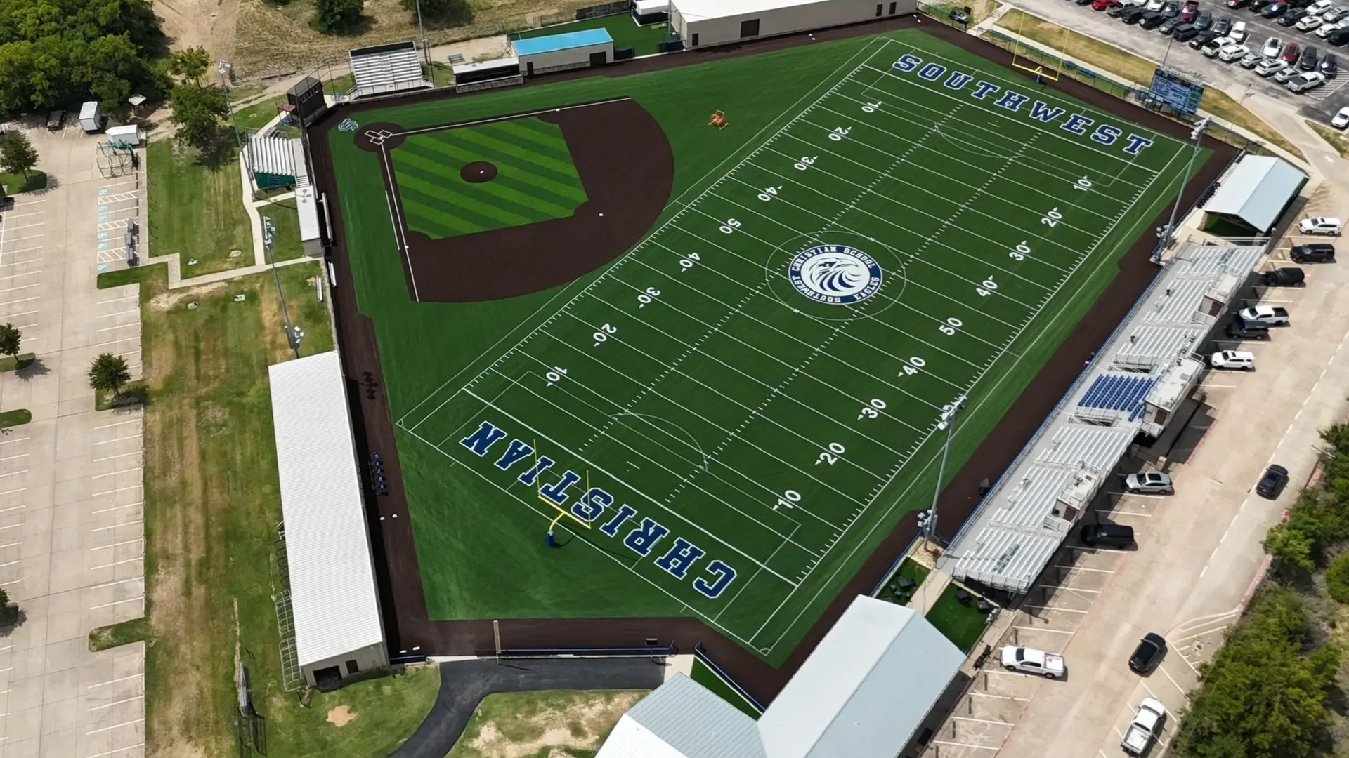 Aerial view of a green football field with white yard lines and a brown baseball diamond on the left