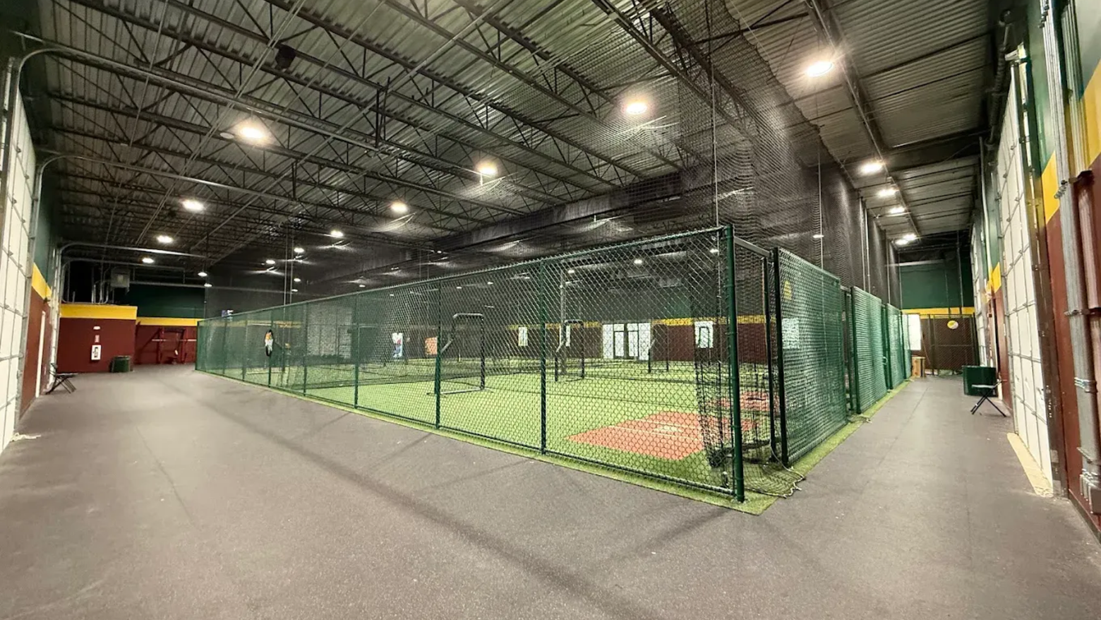 Indoor sports facility with batting cages, green netting, and a wide walkway under bright lights