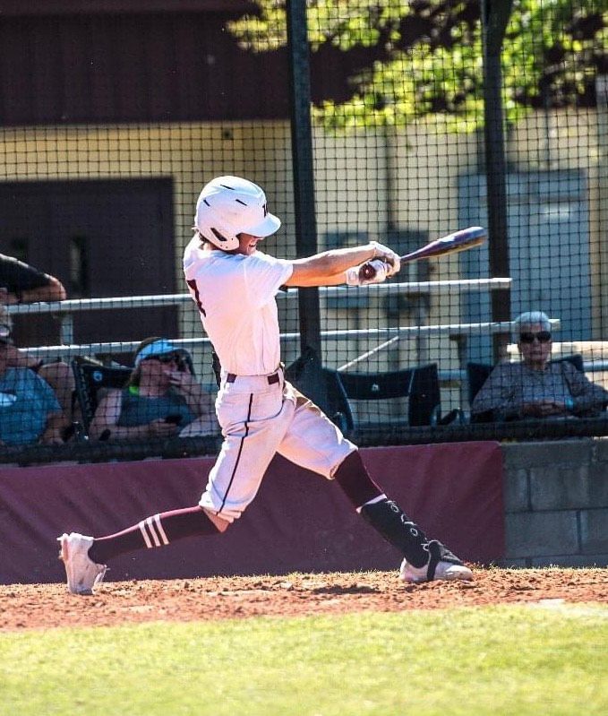 Baseball batter in white uniform swinging at a pitch on a field, with catcher and spectators in the background