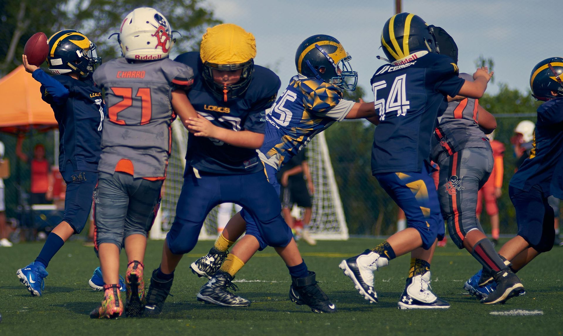 Youth football players in blue and gray uniforms collide during a tackle on a grassy field.