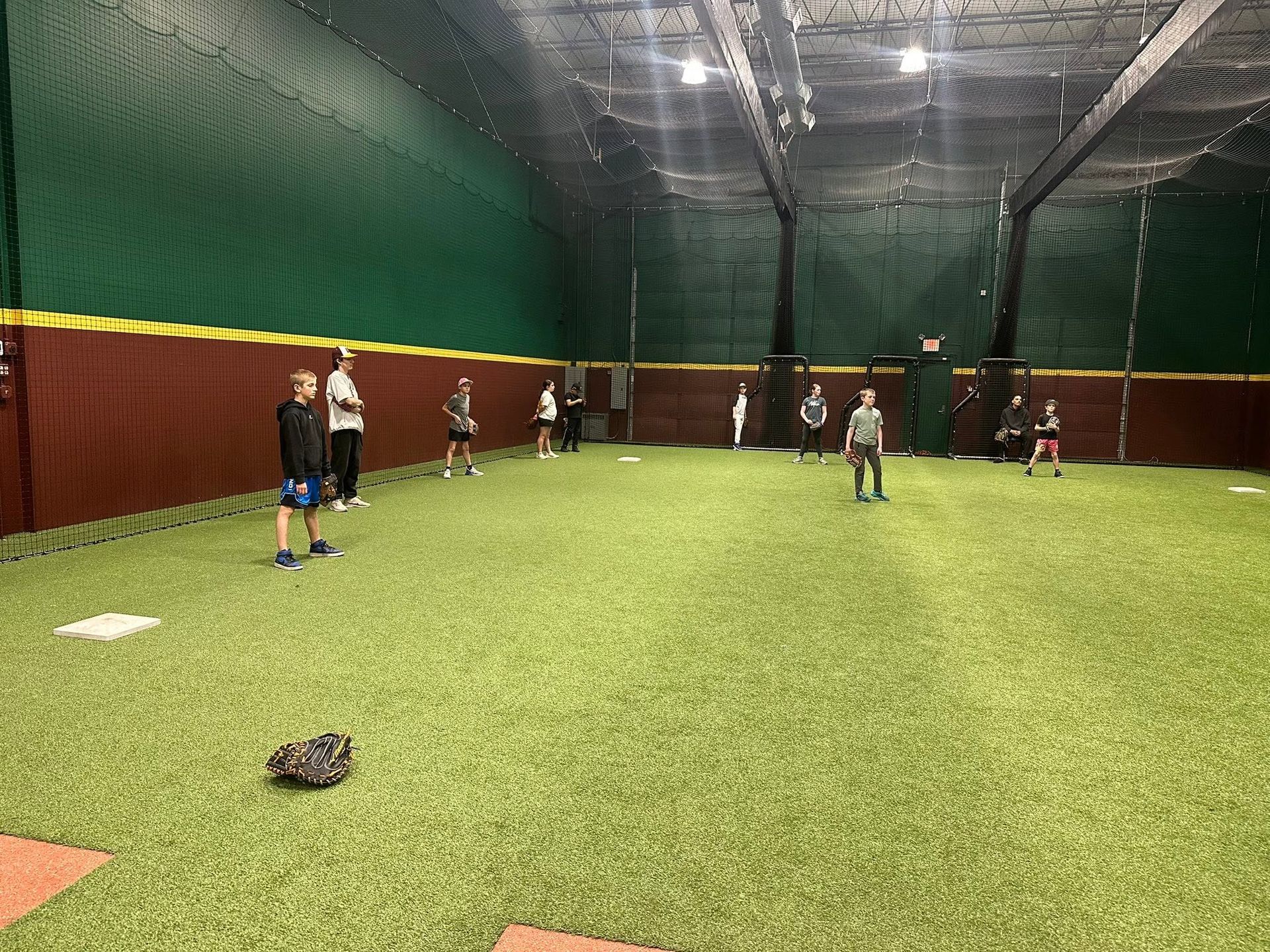 People standing on indoor turf in a large netted practice facility, with baseballs scattered around.