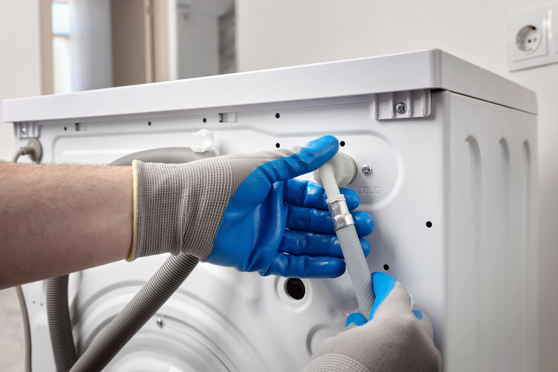 Gloved hands plug a white cable into the back of a washing machine.