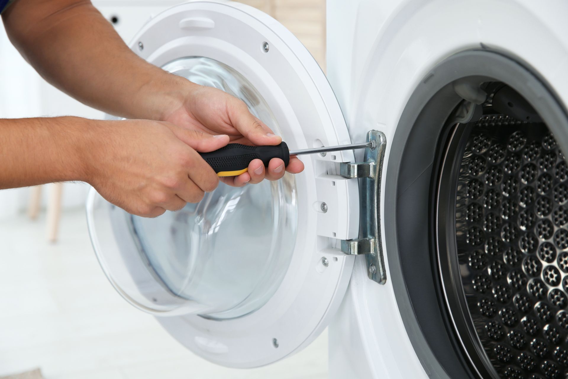 Hands using a screwdriver to repair the hinge of a front-loading washing machine.