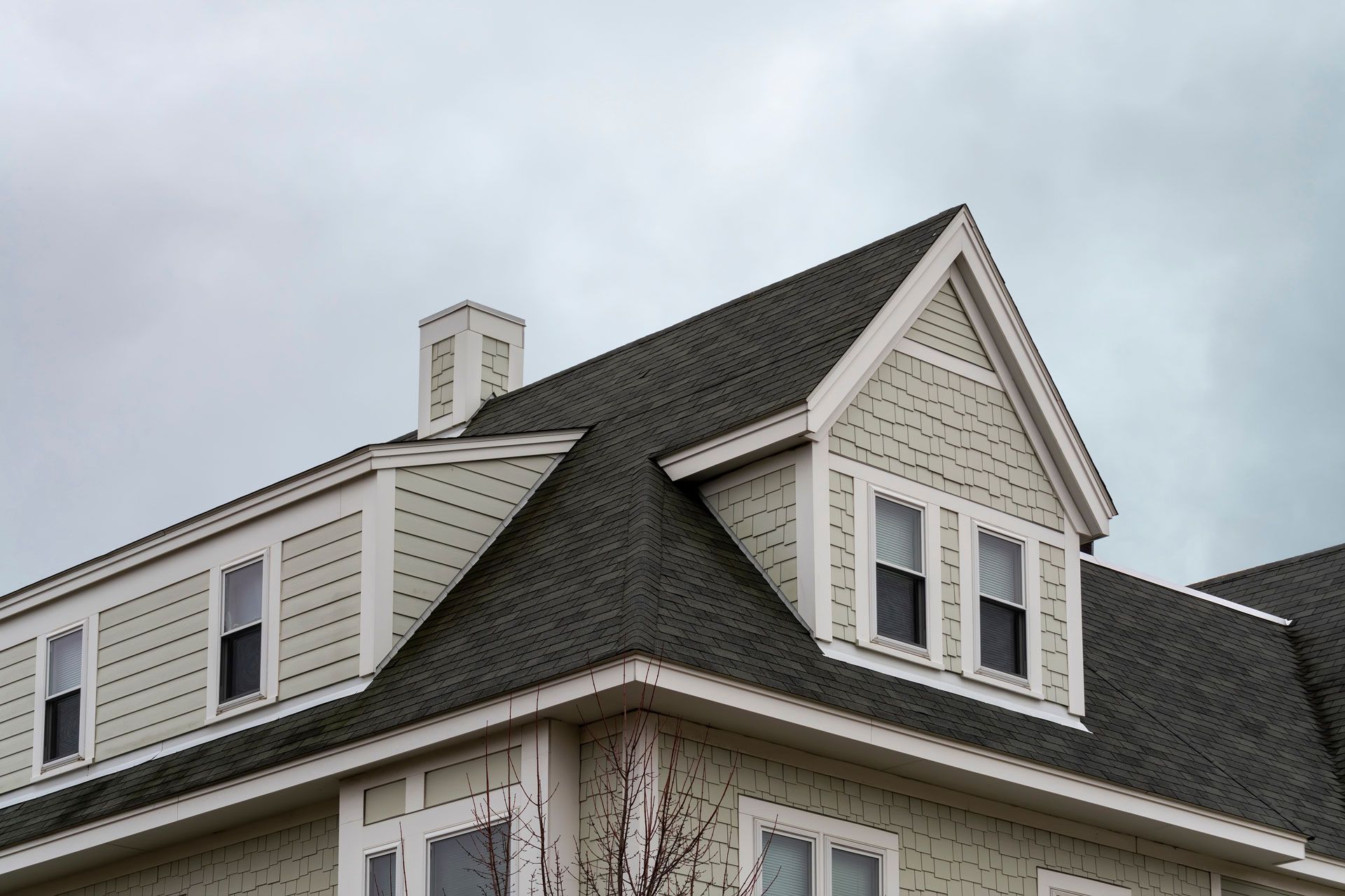 A light green house roof with dark gray shingles, a chimney, and dormer windows against a cloudy sky.
