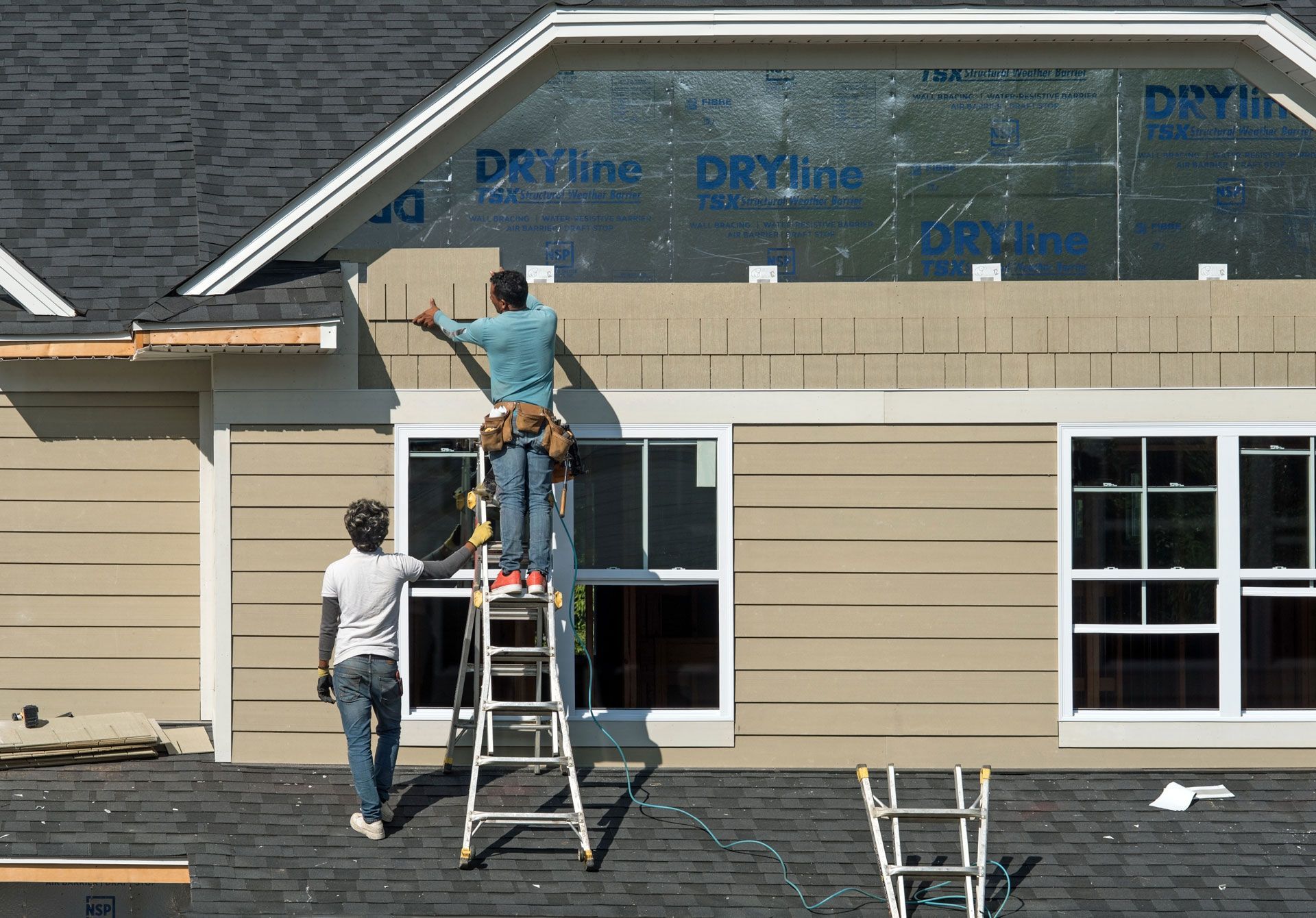 Two construction workers install siding on a house, one on a ladder, tan siding and blue wrap visible.
