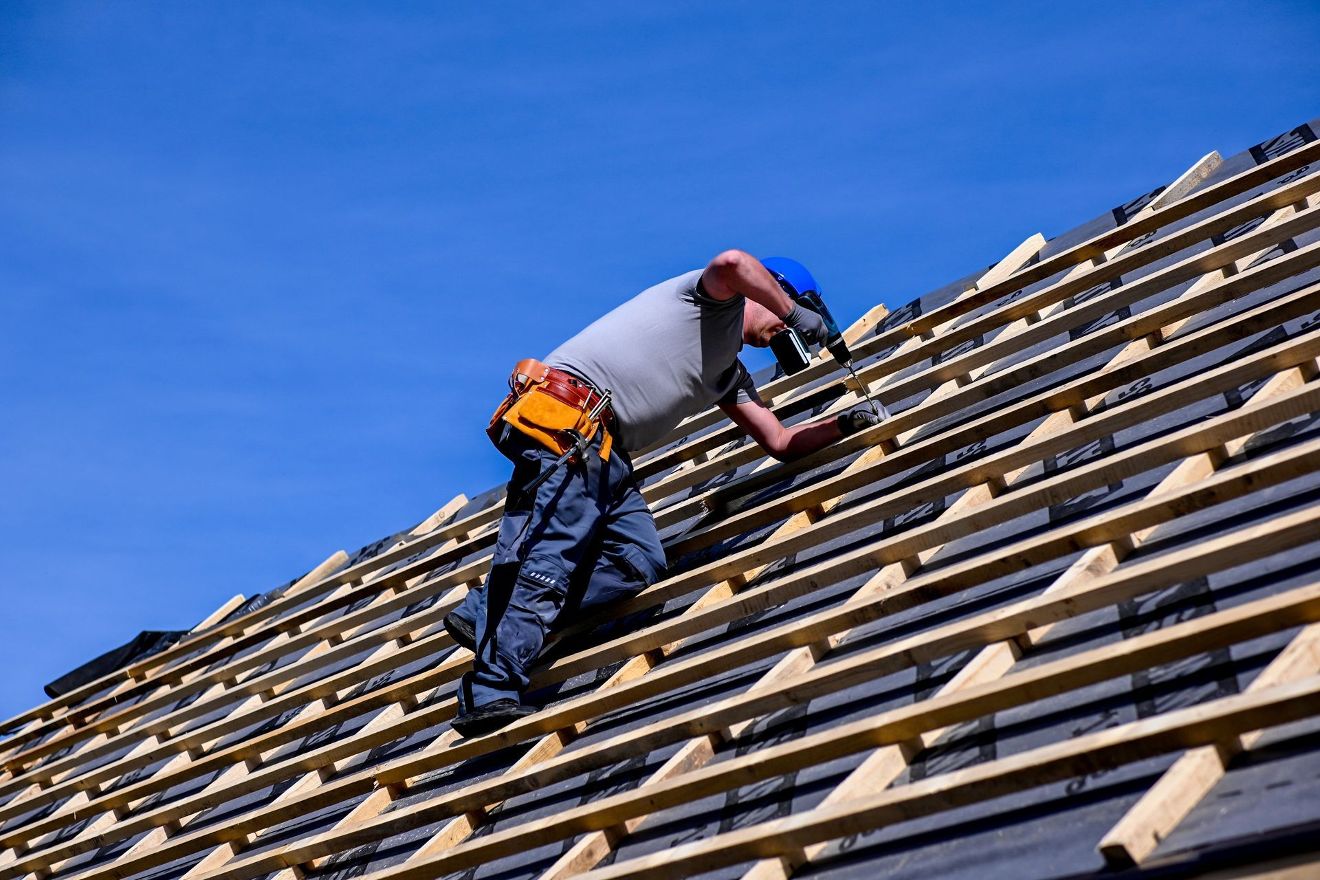 Roofer in a blue hardhat works on a roof under a blue sky, installing shingles.