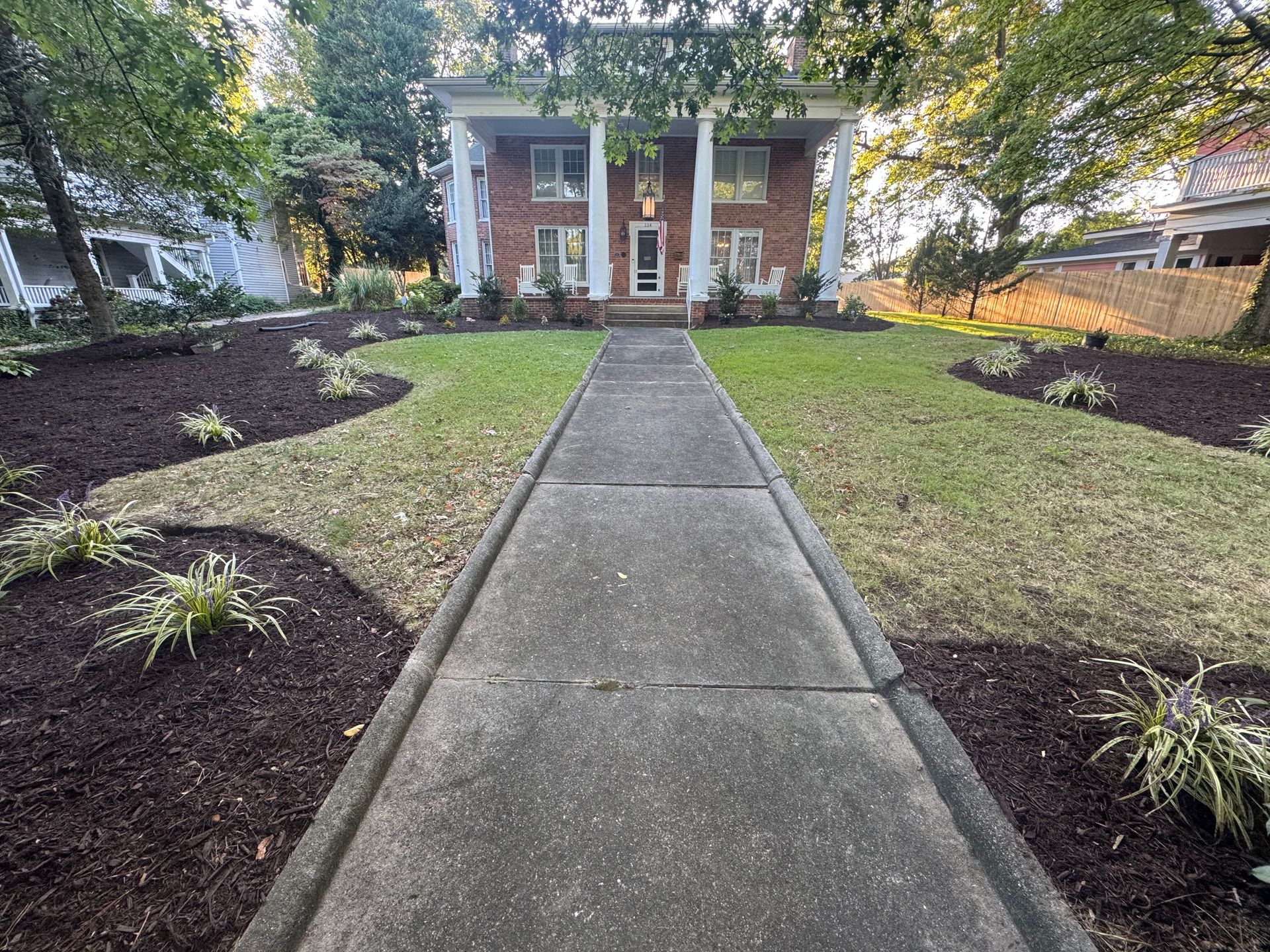 Front walkway leading to a red-brick house with white columns, bordered by mulched beds and green lawn.