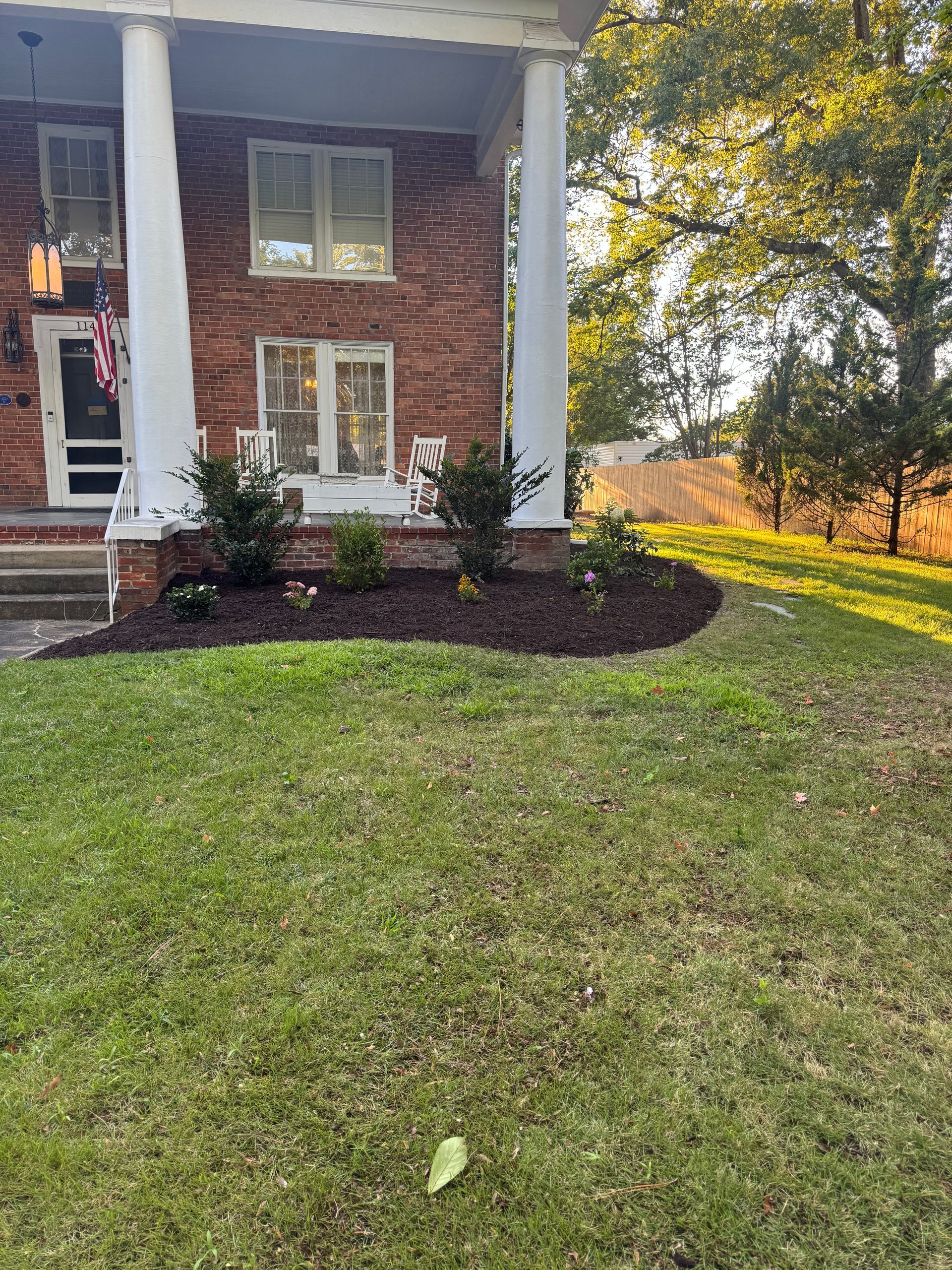 Front lawn beside a brick house with a mulched garden bed and trees in autumn light