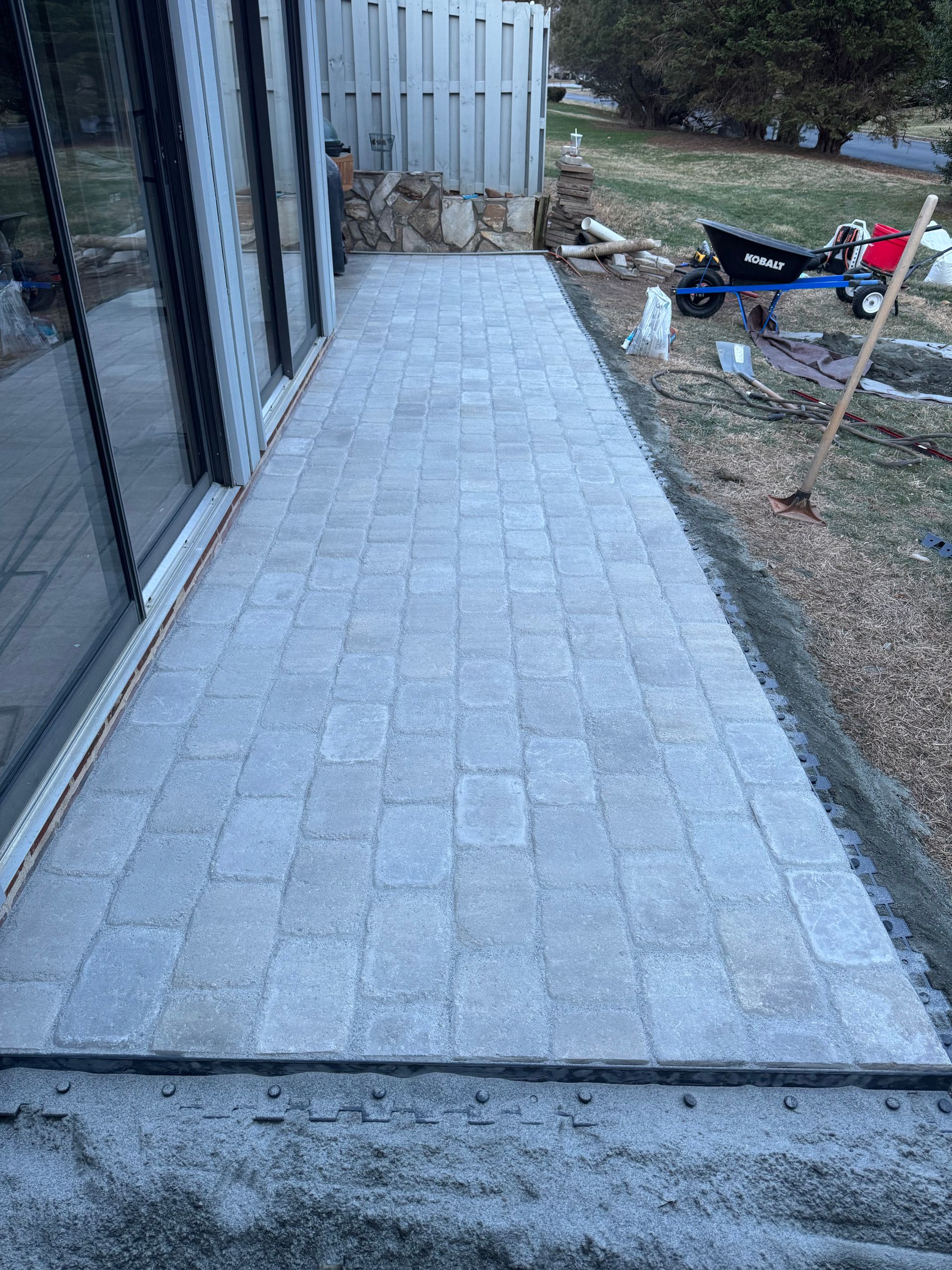 Stone paver walkway alongside a building, leading to a small yard with scattered tools and materials.