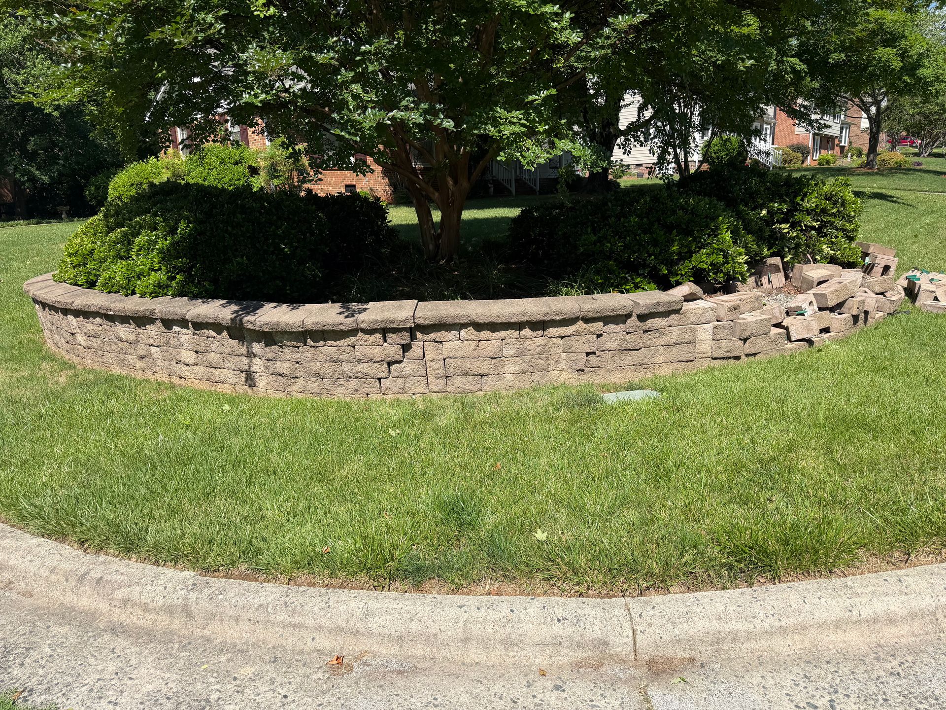 Curved stone retaining wall around a landscaped island with trees and shrubs beside a grassy lawn and sidewalk