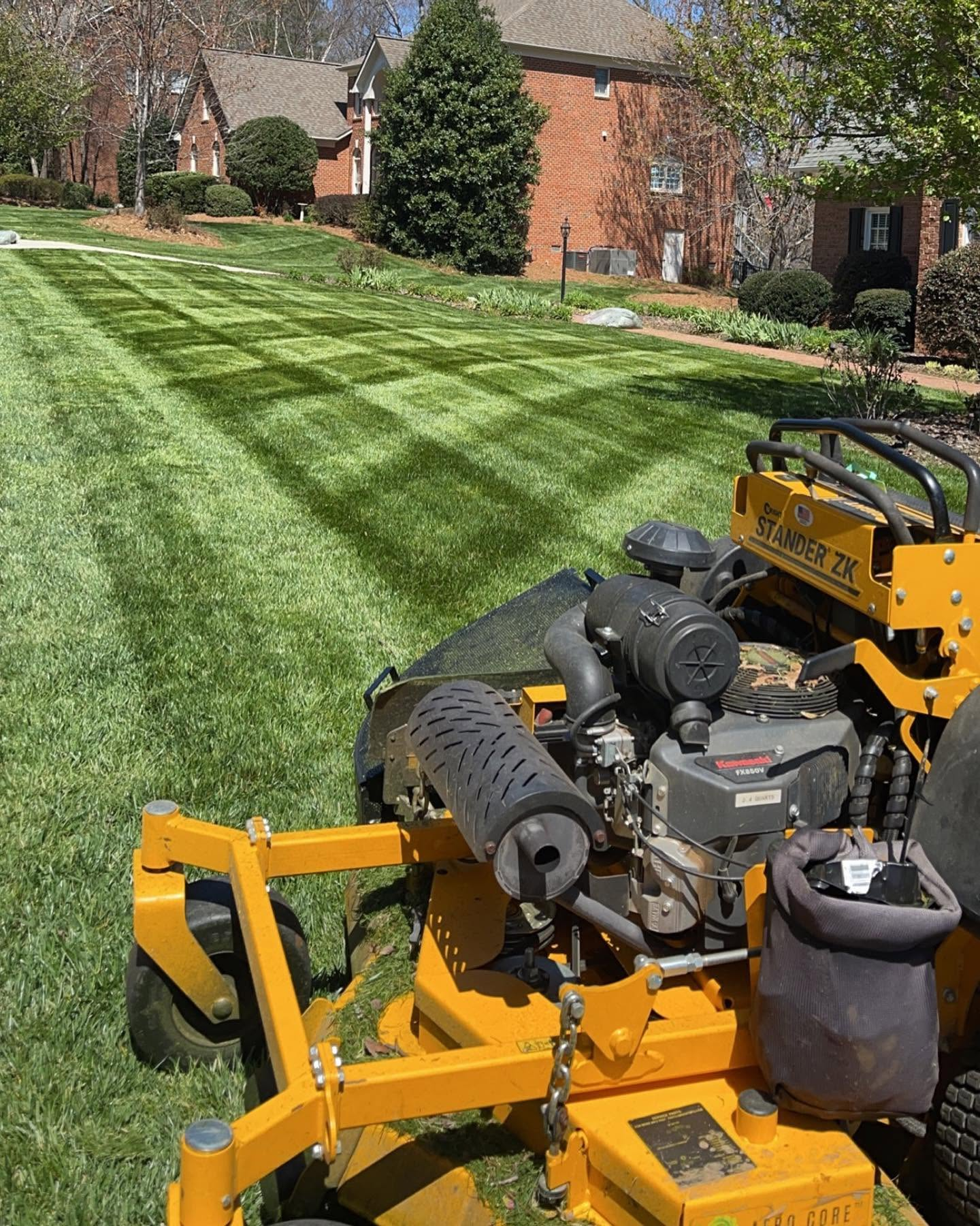 Lawn mower cutting striped grass in front of brick houses on a sunny suburban street
