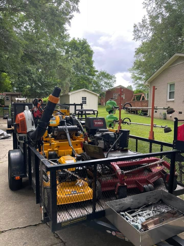 Utility trailer loaded with tools and equipment in a suburban driveway, with bins and a yellow machine visible.