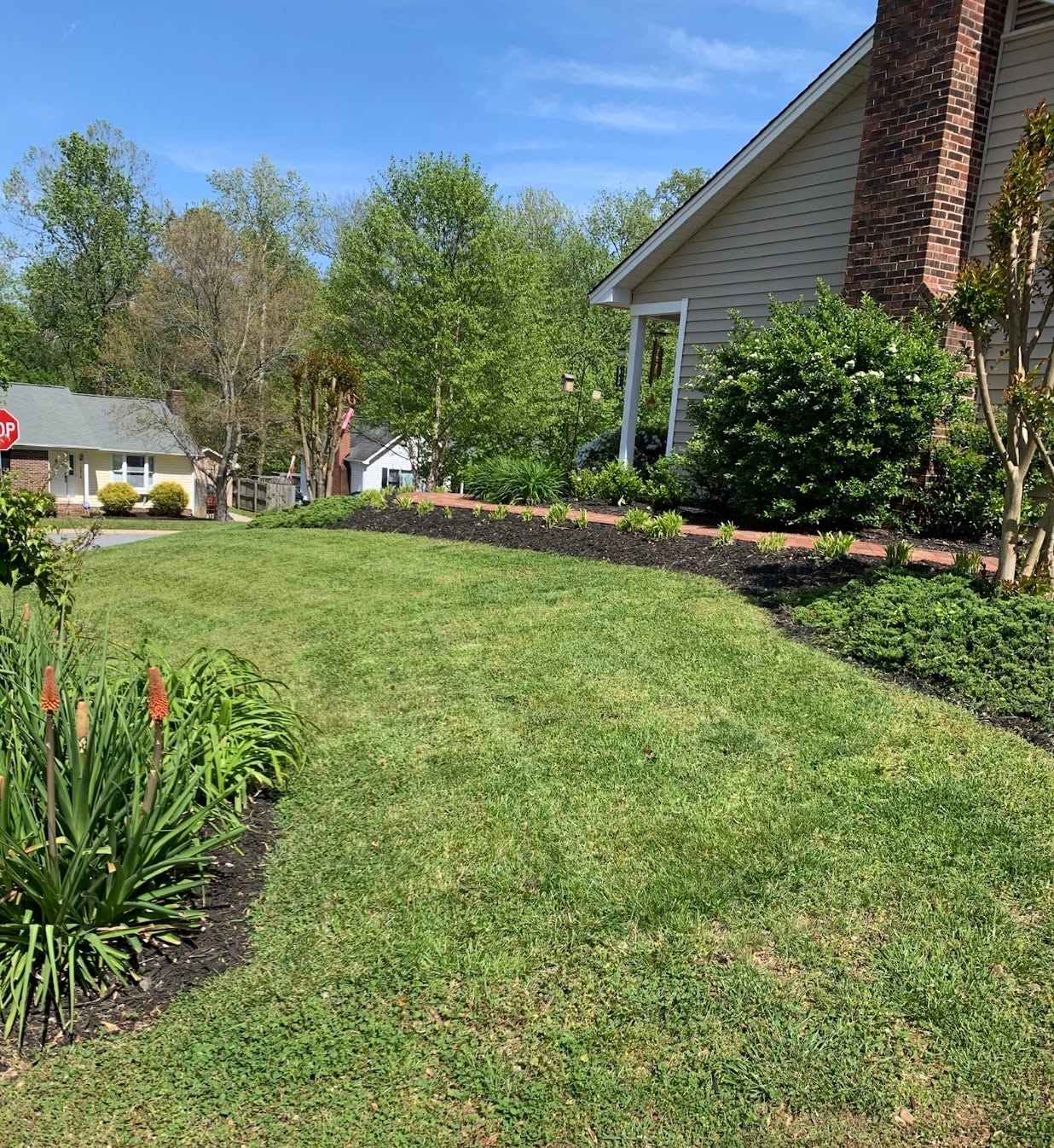 Front yard with neatly mowed green lawn, flower beds, and a brick house under a sunny blue sky