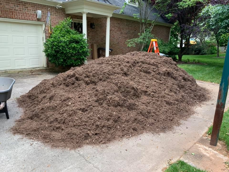 Large pile of brown dirt on a driveway beside a brick house and garage, with a wheelbarrow nearby.