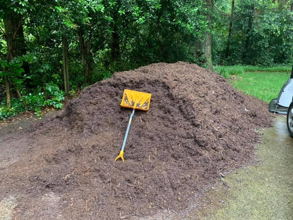 Large mulch pile beside a driveway, with a yellow rake leaning against it near a wooded yard.
