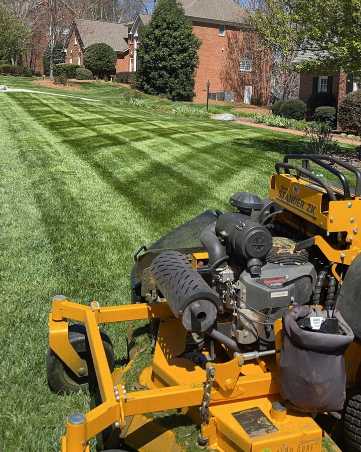 Lawn mower cutting striped grass in front of brick houses on a sunny suburban street