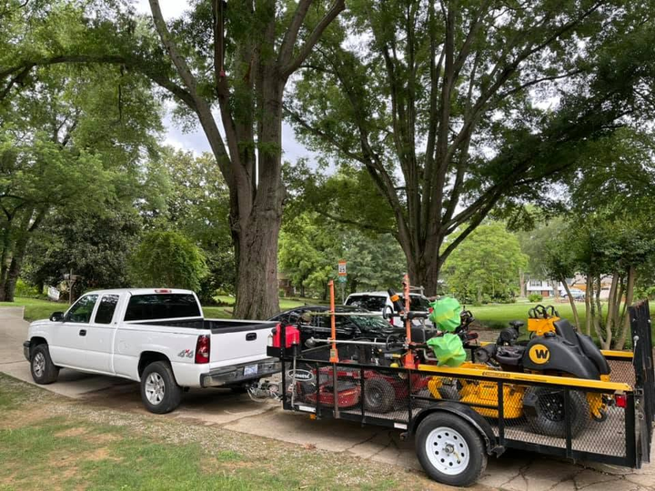 White pickup truck towing a trailer full of landscaping equipment in a grassy park with trees.