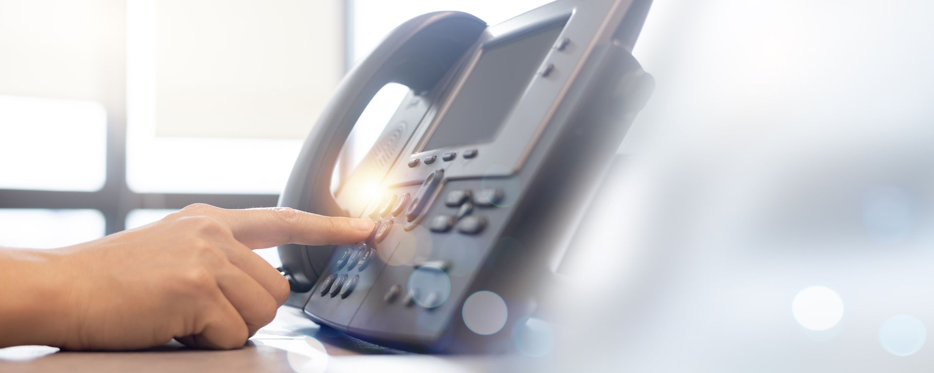 A person's finger presses a button on a black office phone. Bright light shines from behind.