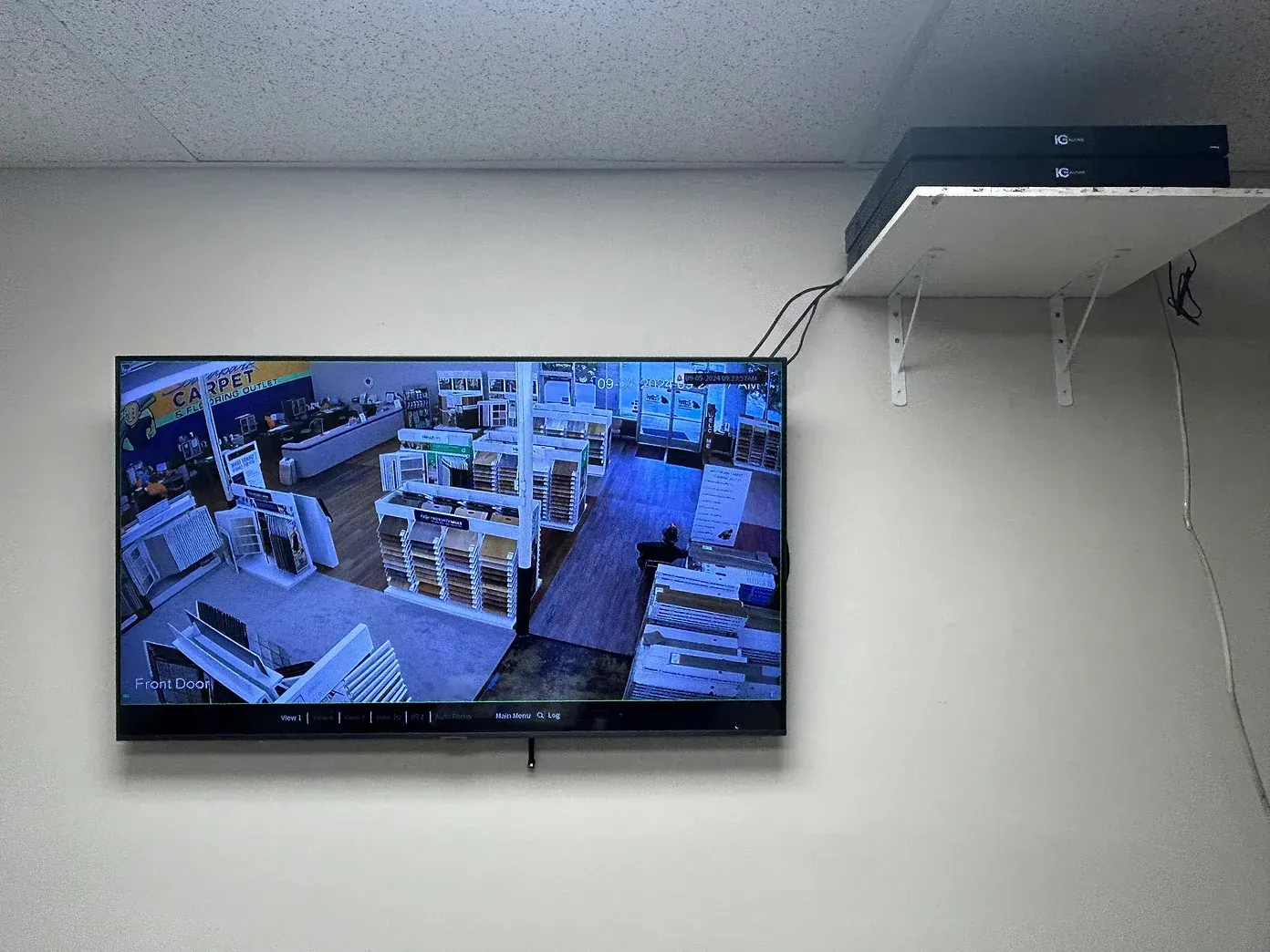 TV screen displaying store security camera footage, mounted on a white wall. Shelf above holds a black DVR.