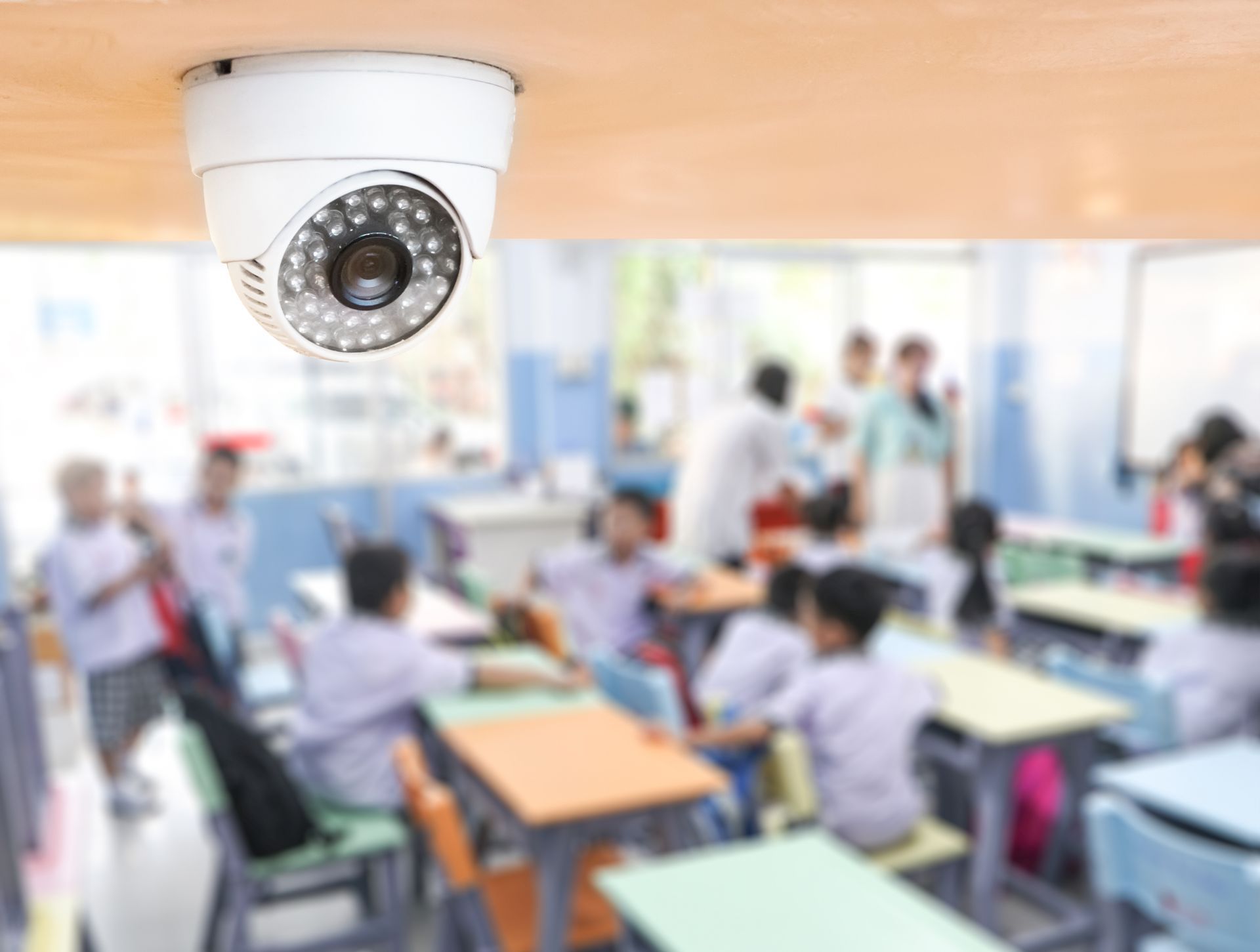 A security camera is hanging from the ceiling of a classroom filled with children.